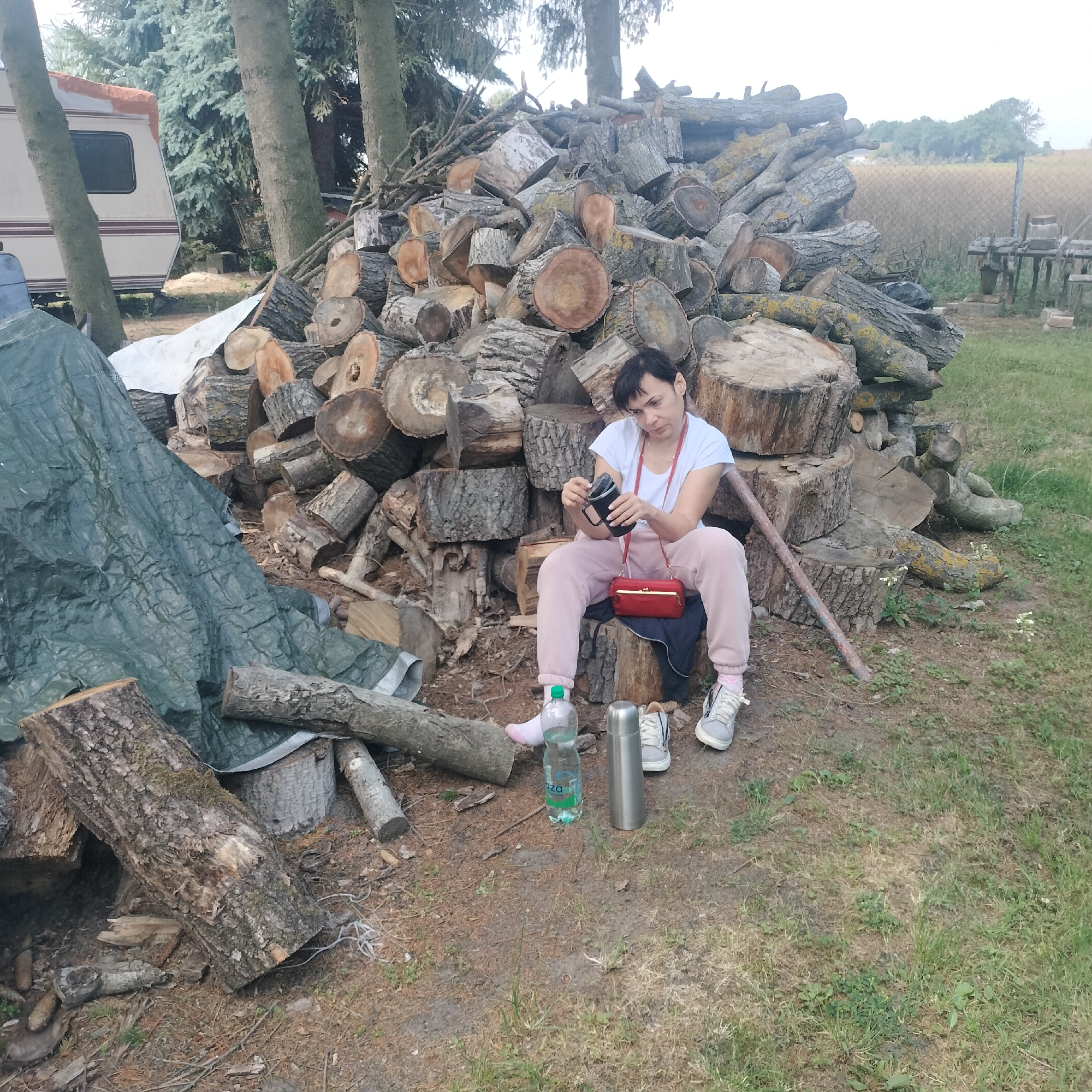 A woman with short dark hair, wearing a white t-shirt, light pink sweatpants, and light pink socks with gray sneakers, sits on a cut log in front of a large pile of chopped wood. She is holding a black object, possibly a camera, in her hands and looking down at it. A red clutch purse with a gold clasp is slung across her body. Next to her are a clear plastic water bottle, a silver thermos, and another pair of gray sneakers. The scene is outdoors, with trees and grass in the background. A portion of a camper trailer is visible on the left side of the image. This location is identified as Witków, Poland.