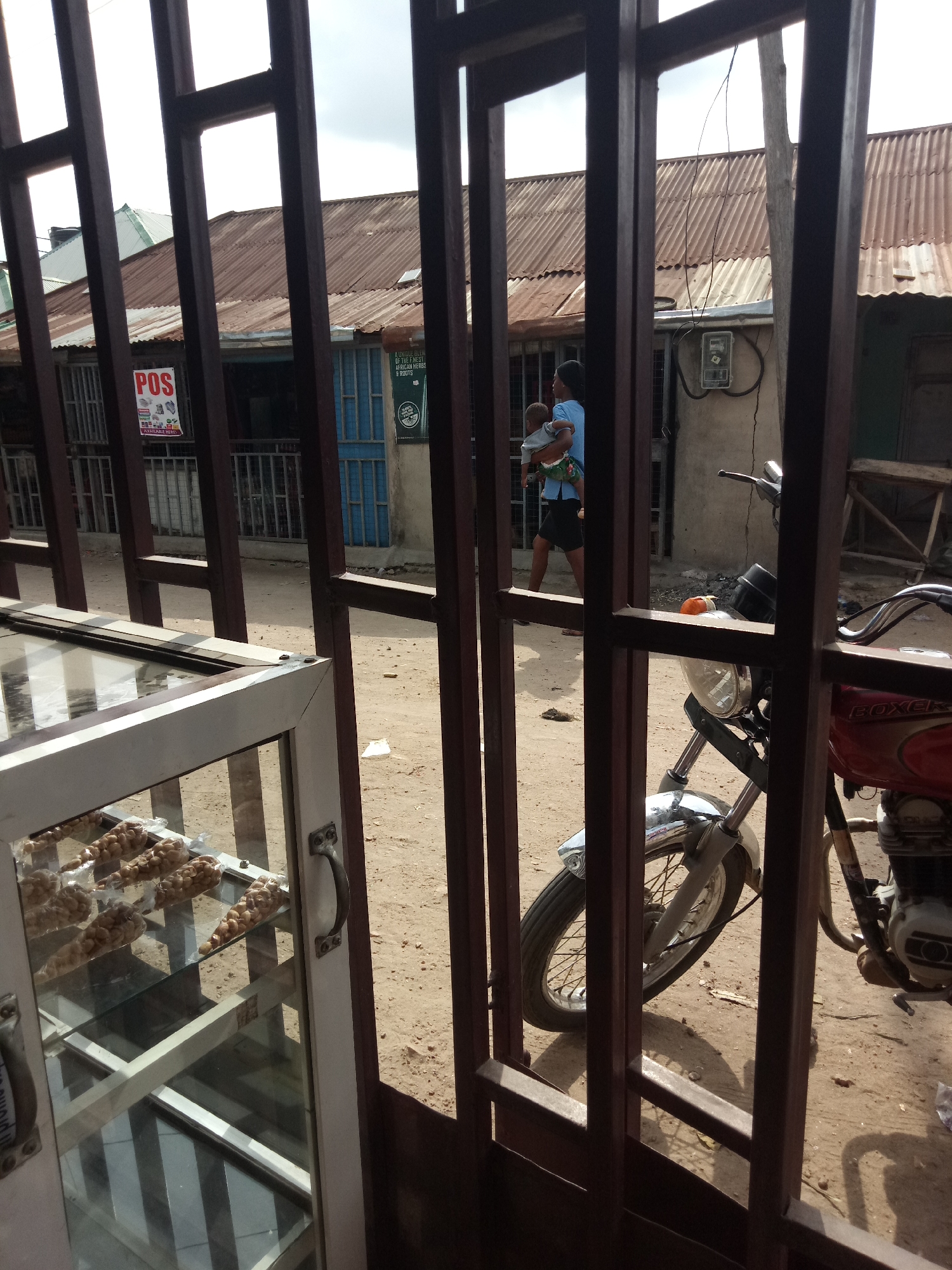 In an outdoor, gritty urban environment, possibly a market or street, a person carries a child while walking past corrugated metal-roofed buildings. A motorcycle is parked on the dirt ground in front of one of the buildings, partially obscured by a metal gate. The gate's dark brown bars are in the foreground, creating a visual barrier. To the left, a glass display case filled with packaged snacks is visible. The overall scene depicts a busy, perhaps impoverished, street scene with elements of commerce and daily life.