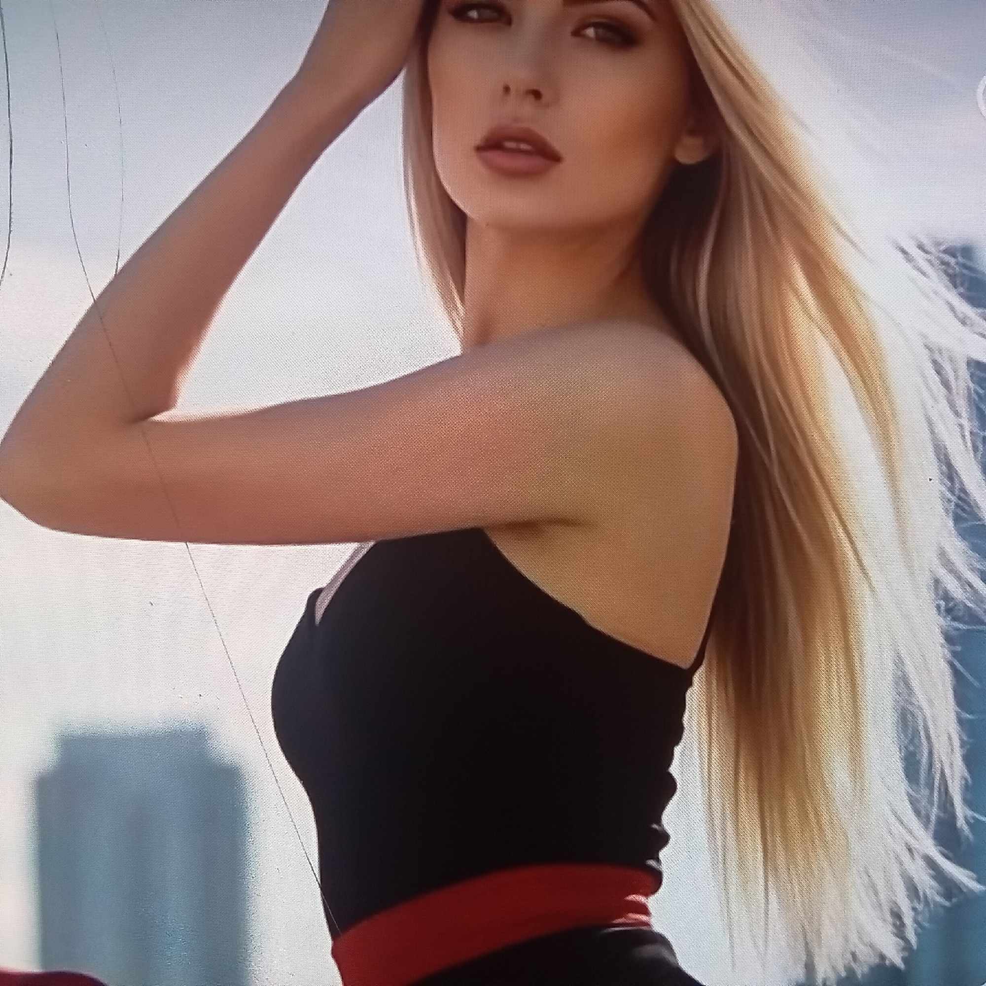 The image shows a close-up of a blonde woman with fair skin and full lips, wearing a black dress with a red sash. Her hair is long and flowing, and her left arm is raised, resting on her head. She is looking towards the viewer with a neutral expression.
The background is a blurred cityscape, suggesting an urban setting. The lighting indicates it is daytime, likely with bright sunlight, as evidenced by the highlights on her arm and hair. The presence of buildings in the background and the overall bright atmosphere suggest an outdoor location, possibly a rooftop or a high vantage point in Almaty, Kazakhstan. The image has a somewhat grainy quality, with some faint lines visible across the surface, possibly from a screen or a damaged print. There is no visible text in the image.