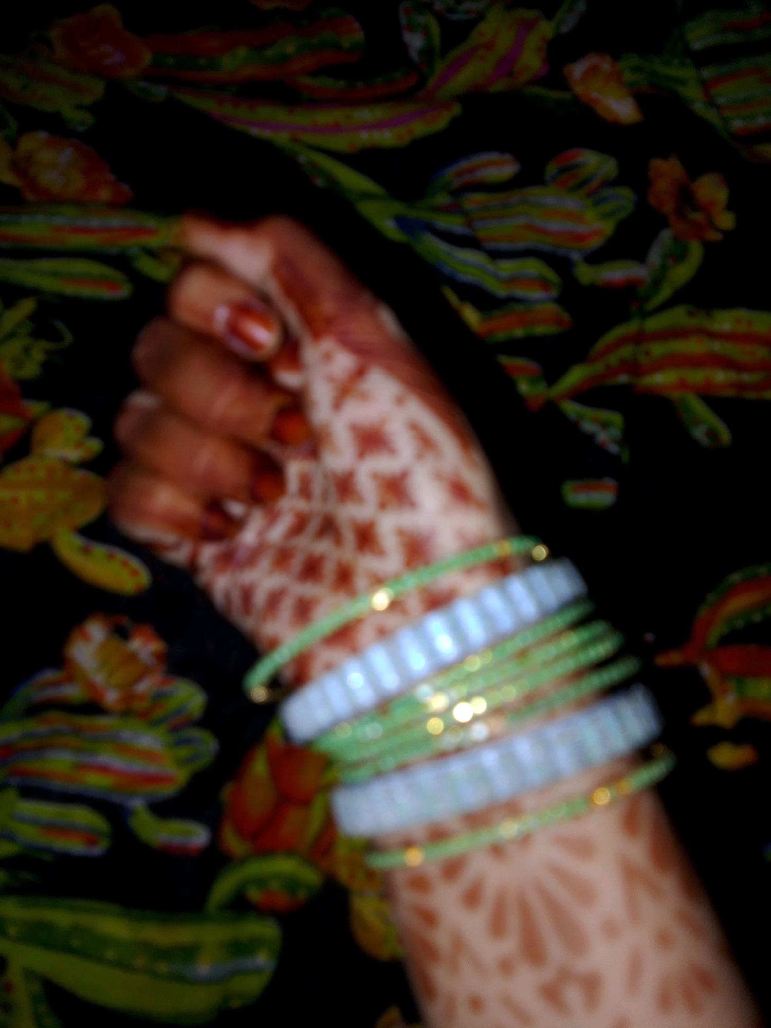 A close-up, blurred shot shows a woman's henna-decorated hand and wrist adorned with several bangles. The bangles are a mix of light blue and green, with some featuring small golden accents. The henna pattern on the hand is intricate, resembling floral or geometric designs. The background is a dark fabric with a colorful, abstract floral print. The image quality is poor, with low resolution and a lack of sharp focus, making it difficult to discern finer details. The setting is not identifiable as being in the city of Thane, India.