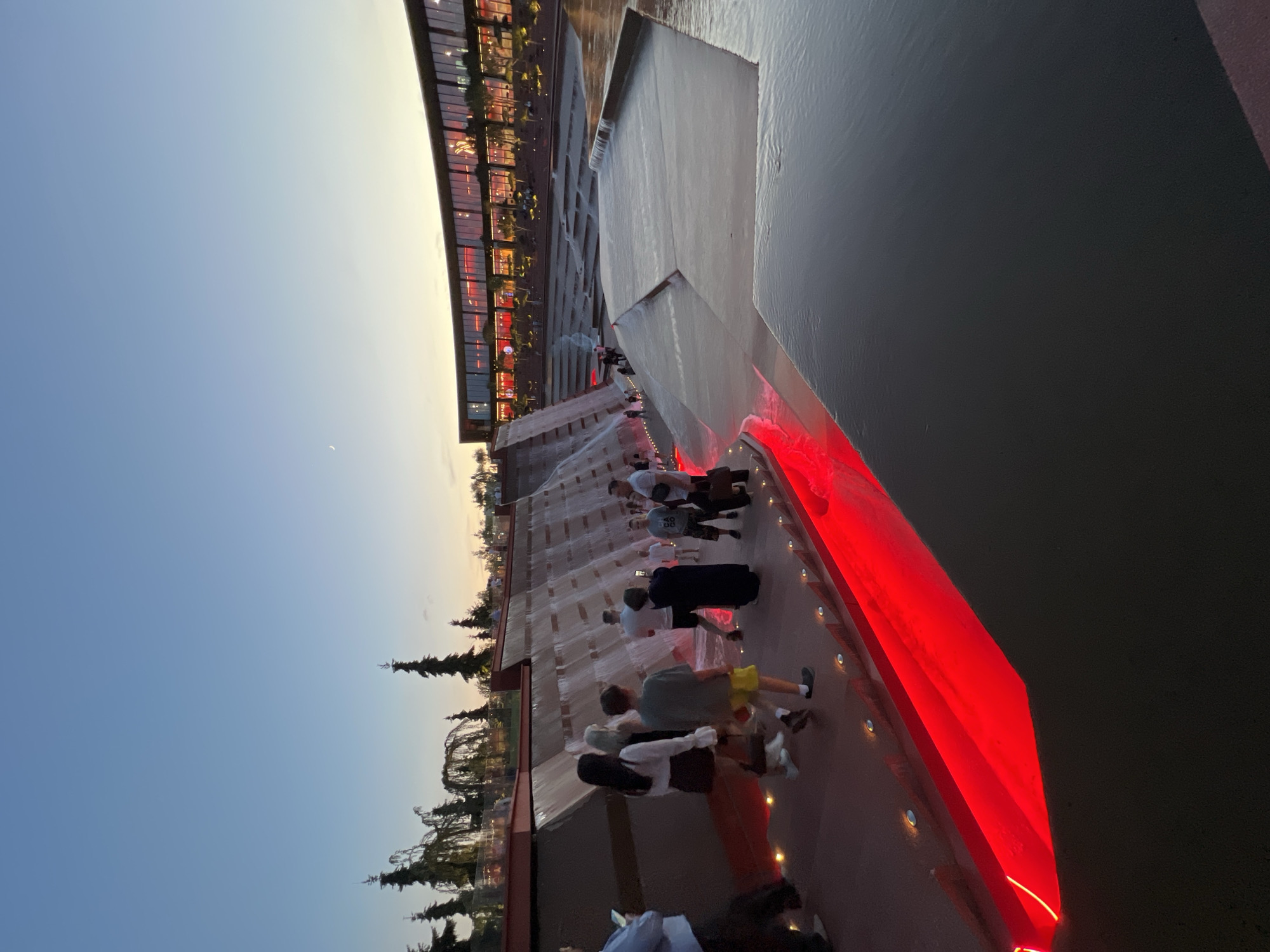 This image captures a group of people gathered at an outdoor modern architectural installation featuring a large, tiered water feature. The water cascades down the multi-level structure, creating a dynamic visual. A prominent, bright red light illuminates one side of the water feature, casting a vibrant glow on the water and the surrounding concrete surfaces. The architecture appears to be made of concrete and glass, with a curved building visible in the background, showcasing illuminated windows and what looks like shops or restaurants.
Several individuals are seen walking along the pathways and steps of the water feature, observing the spectacle. Some are casually dressed, suggesting a leisurely outing. The time of day appears to be dusk or early evening, as evidenced by the clear, pale blue sky with a hint of twilight. A faint crescent moon is visible in the sky. The overall atmosphere seems relaxed and enjoyable, with people appreciating the unique design and the interplay of light and water.
The scene is set in an urban or developed area, with evergreen trees and hints of landscaping suggesting a public park or a modern plaza in Krasnodar, Russia. The lighting from the red element and the illuminated building indicates that the area is designed for evening or nighttime enjoyment. There is no visible text in the image.