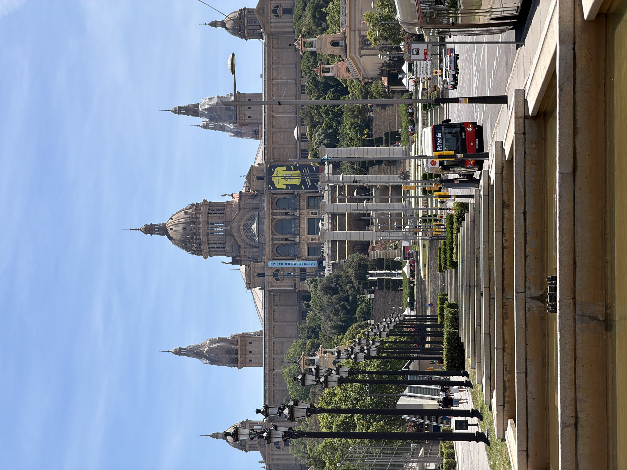The image shows a wide, multi-tiered fountain in the foreground with a grand building in the background. The building features multiple domes and towers, indicative of a significant architectural landmark. A wide set of stairs ascends towards the building, flanked by rows of lampposts. A red bus with "Ps. Maritim" displayed on its front travels on a road to the right of the stairs. Several cars and a tram are also visible on the road. The sky is clear and blue. The overall setting suggests a public space in Barcelona, likely the area in front of the Palau Nacional, which houses the Museu Nacional d'Art de Catalunya, on Montjuïc hill. The presence of the bus route name "Ps. Maritim" is consistent with Barcelona's public transport system.