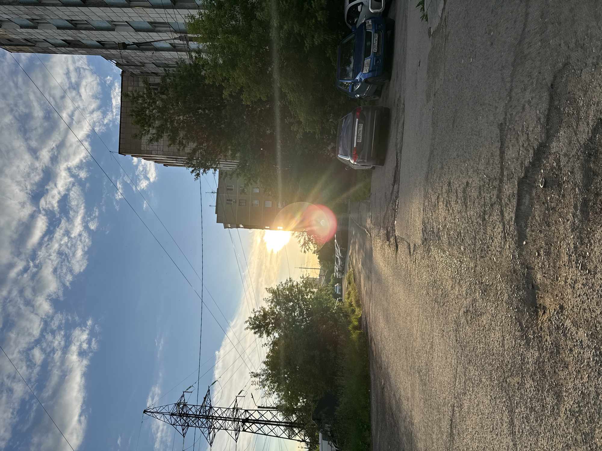 The image depicts a street scene in the city of Perm, Russia, during a sunset. The sun, low on the horizon, creates a strong lens flare effect on the right side of the frame, illuminating the sky with warm hues and casting long shadows. Dominating the left side of the image is a tall, industrial-looking electricity pylon with multiple crossbars and wires extending across the sky. Adjacent to the pylon is a dense cluster of green trees and foliage, partially obscuring the view behind it.
In the background, several multi-story apartment buildings are visible. The building closest to the right edge of the frame has a facade with a grid-like pattern of light blue and grey panels. Further back, another apartment building, appearing older and possibly made of concrete, is visible. A line of parked cars is situated along the right side of the street, including a dark sedan with a license plate reading "0887ME 159" and a blue SUV with a license plate "P207VB 159". A white car is partially visible to the right of the blue SUV.
The street itself appears to be paved with asphalt and shows signs of wear, with visible cracks and patches. The lighting suggests it is late afternoon or early evening. The sky is blue with scattered white clouds, some of which are illuminated by the setting sun. No people are visible in the image.