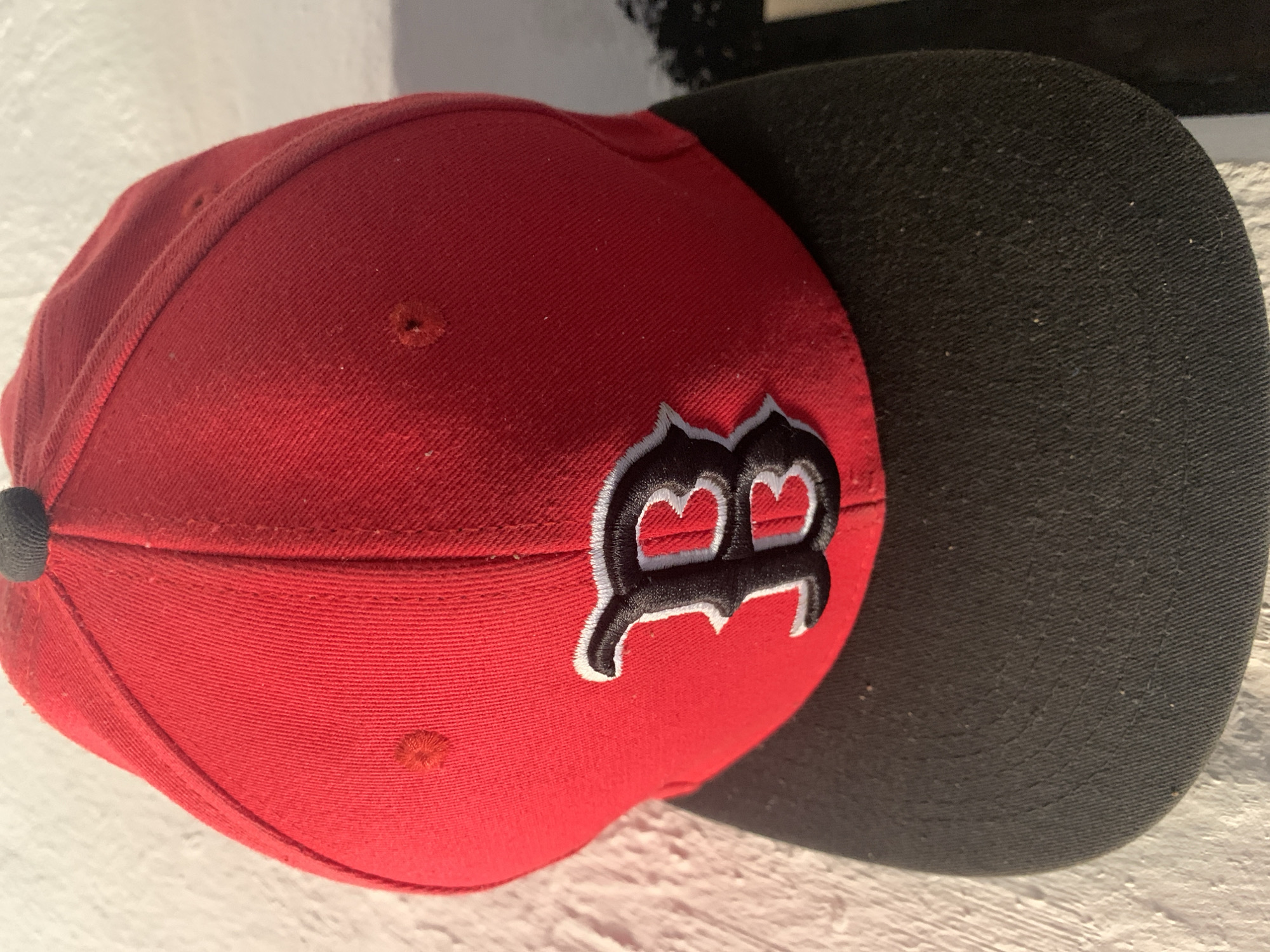 The image features a close-up of a red and black baseball cap, positioned on its side against a light-colored, textured background. The main body of the cap is a vibrant red, made of a visible fabric texture, and includes small, circular ventilation eyelets, two of which are clearly visible. A prominent, stylized letter "B" is embroidered on the front panel of the cap. This "B" is black with a distinct white or light gray outline, consistent with the logo of the Boston Red Sox baseball team.
The brim of the cap is solid black, also made of a fabric material, and shows some minor dust or lint on its surface, indicating casual use. A small black button-like fixture is visible at the very top of the cap. The lighting in the image appears to be natural daylight, coming from the right, which creates soft shadows under the brim and along the contours of the cap. The overall impression is one of a well-used, iconic piece of sports apparel. No people or specific location cues are visible, keeping the focus entirely on the cap.