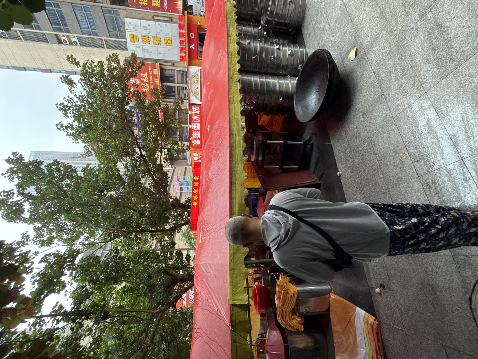 The image depicts a street scene in Haizhu District, China, where a vendor appears to be preparing for or operating a food stall. A person wearing a grey hooded sweatshirt and patterned pants stands facing away from the camera, looking towards the stall. The stall itself is covered by a large red and green tarp, suggesting it might be a temporary setup.
Several large, metallic steamer baskets are stacked neatly to the right of the stall. In front of the stall, a large, dark wok-like cooking vessel is placed on the ground, possibly for immediate use or storage. Various containers, including red bowls and metal pots, are visible under the tarp, along with neatly folded yellow cloths. The ground is paved with grey square tiles, and a few fallen leaves are scattered on the pavement. In the background, tall buildings and trees suggest an urban environment. Signs with Chinese characters are visible on the buildings, indicating the commercial nature of the area. The sky appears overcast, and there are water droplets on the tarp, suggesting it might have recently rained or is still drizzling.