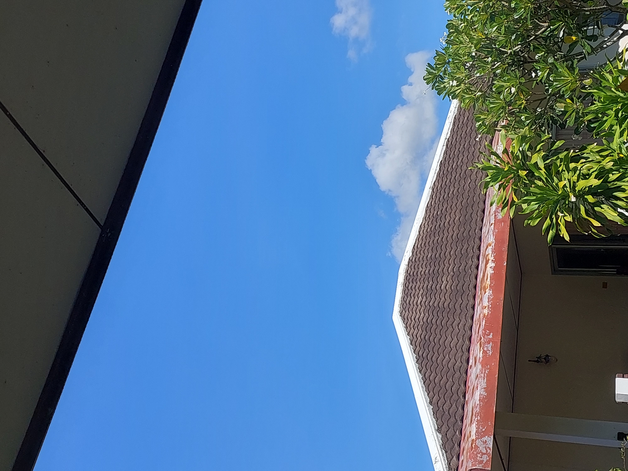 The image depicts a clear blue sky over a residential building. In the foreground, a portion of a dark-colored overhang is visible. The roof of the building is covered with brown, curved tiles, and a white fascia board runs along its edge. Below the roof, a red awning or trim shows signs of wear. Green foliage from trees and plants is present in the lower right and mid-right of the frame. The building appears to be a single-story structure with light-colored walls and a visible doorway or window on the right. A hanging light fixture is visible on the exterior wall. The scene is situated in Baan Mae Nam, Thailand, under bright daylight conditions. No people are present in the image.