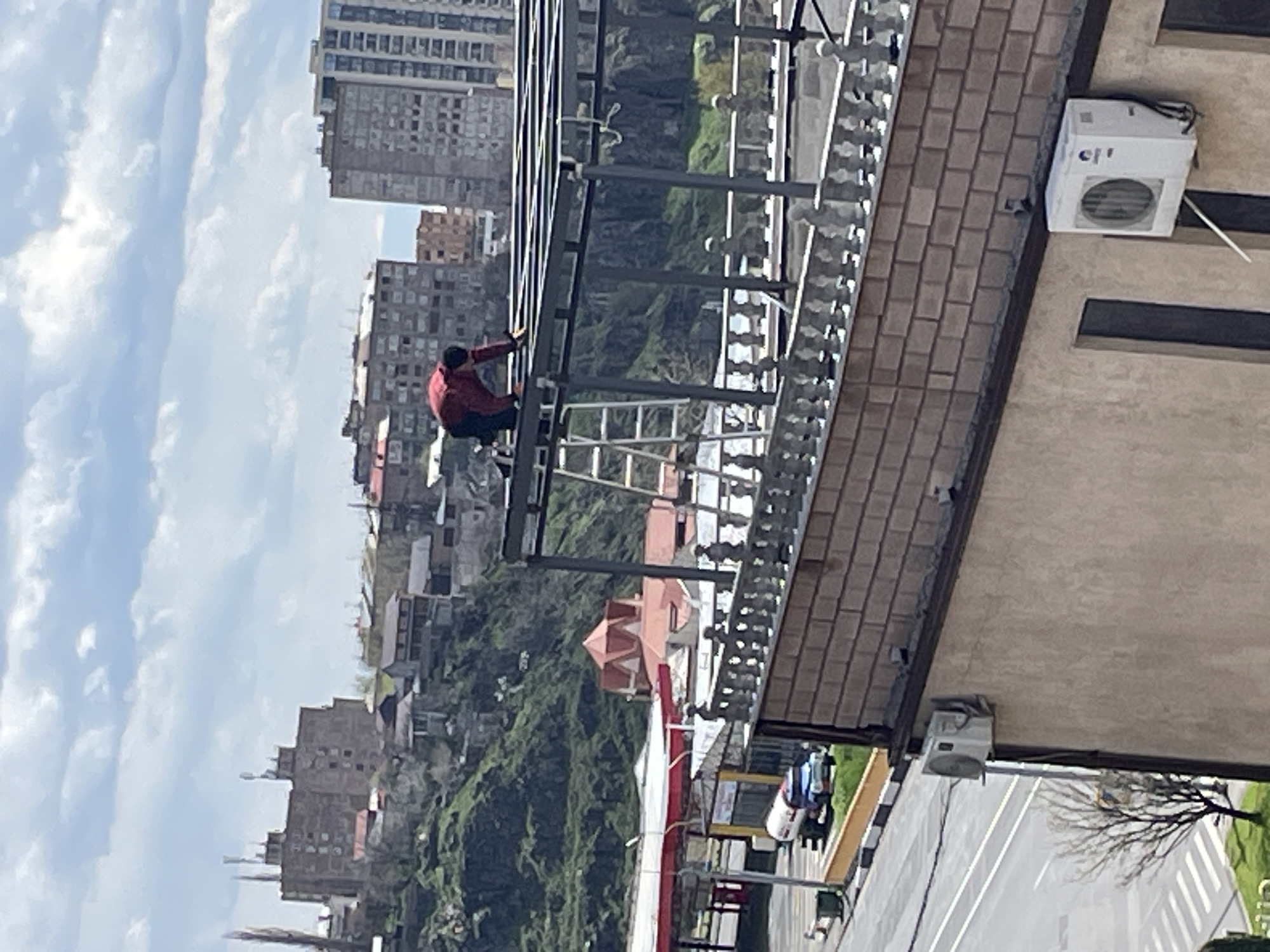 A man is working on a metal structure on a rooftop in Yerevan, Armenia. He is wearing a red jacket and is crouched down, appearing to adjust or secure something. A ladder is positioned nearby, likely used for access. The metal structure overhangs the roof, with a view of buildings, green hills, and streets below. The rooftop has a brick edge, and an air conditioning unit is mounted on the wall. In the background, the sky is partly cloudy. A street and traffic lights are visible below the rooftop.