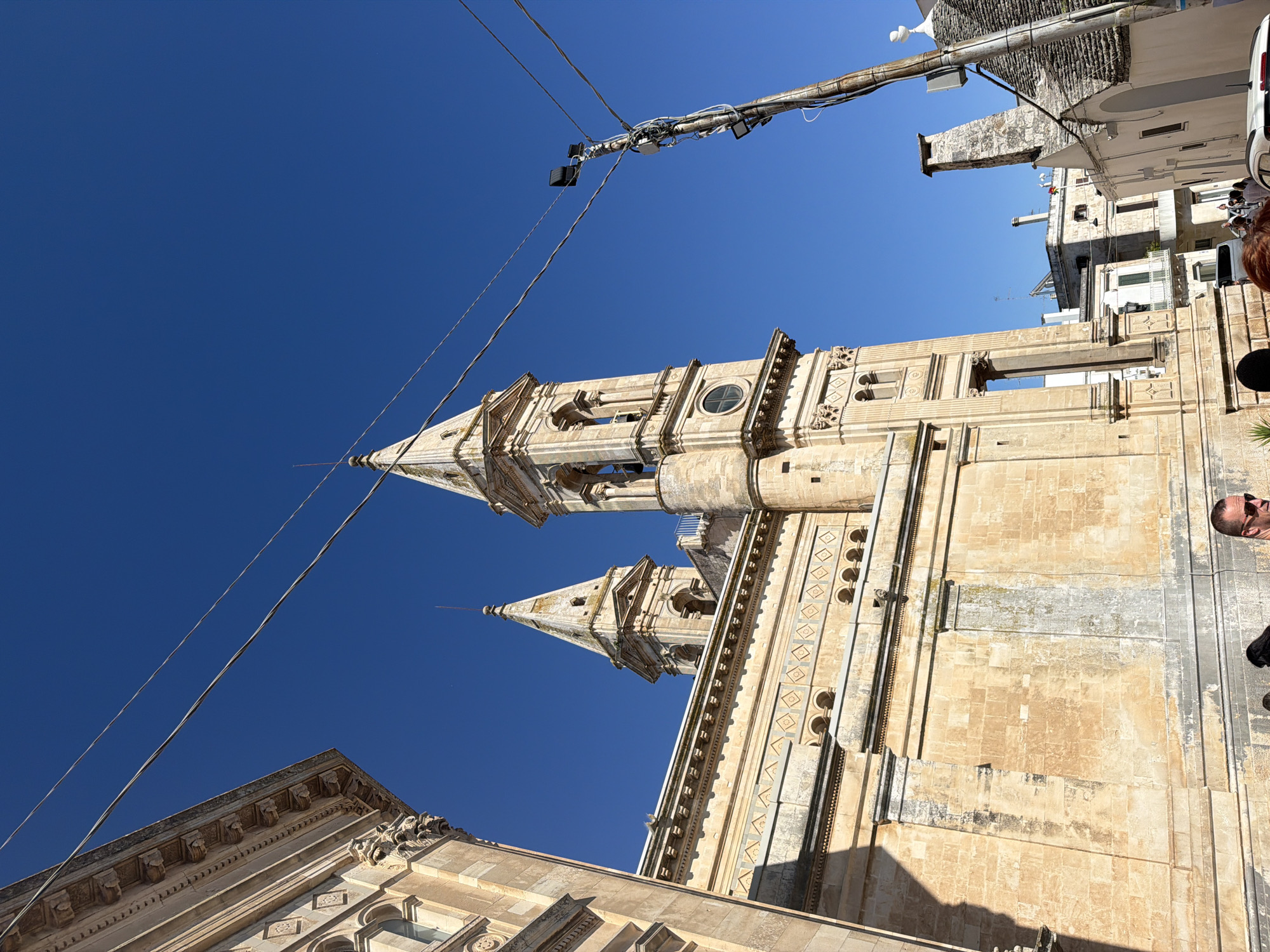 The image shows a clear, bright blue sky over the city of Alberobello, Italy, known for its trulli houses. A large stone building, likely a church or historical monument, dominates the left side of the frame. It features ornate architectural details, including decorative friezes and arched openings. Two bell towers rise from the building, one with a clock face visible.
In the foreground, and slightly to the right, a utility pole with multiple wires and lights extends upwards. In the background, typical buildings of Alberobello, including a trullo with its distinctive conical roof, are visible. The scene is bathed in sunlight, casting shadows. Several people are present, though their features are not clearly discernible due to the distance and angle, with some walking and others standing. The overall impression is a sunny day in a historic Italian town.