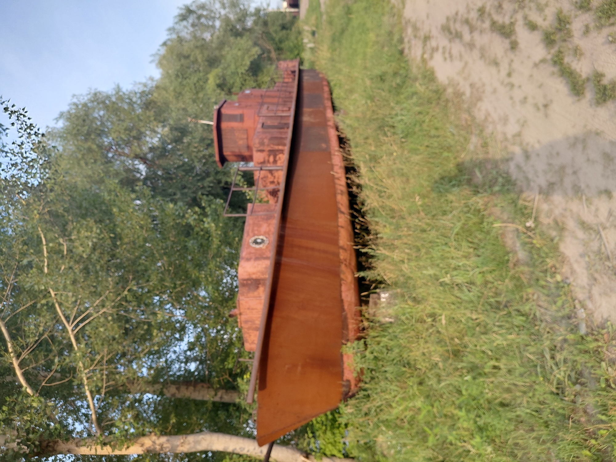 The image depicts a rusty, abandoned barge beached on a grassy embankment next to a dirt path. The barge is old and appears to be made of metal, with significant rust and wear visible. It has a cabin structure at the stern, with windows and railings. The surrounding environment is natural, with trees and overgrown grass. The sky is clear and blue. The scene suggests a forgotten or neglected location, possibly on the outskirts of a city or in a more rural area. Without additional information, it is not possible to determine the specific location in Novosibirsk, Russia.