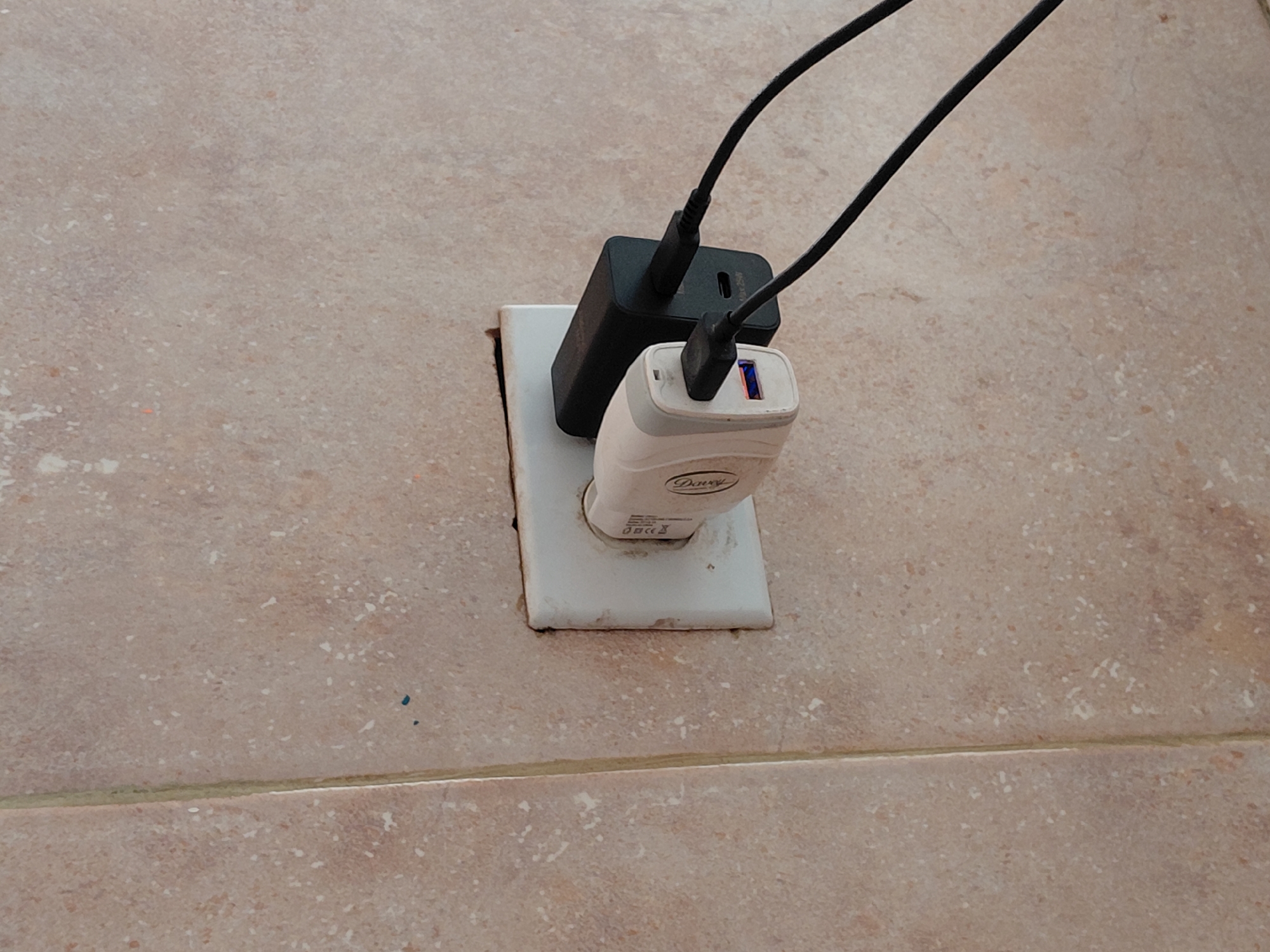 The image displays a close-up view of an electrical wall outlet located on a tiled surface, set in Santa Ana, Venezuela. Two power adapters are plugged into the single outlet. A black, rectangular adapter occupies the upper socket, with faint text "Max 25W" visible on its side. Below it, a white, more rounded adapter is plugged into the lower socket, partially obscuring the black one. This white adapter features the brand "Daway" prominently, along with small regulatory symbols like CE and a crossed-out trash can. Its USB port glows blue, indicating it is active. Two black USB charging cables are plugged into these adapters, extending upwards and out of the frame, suggesting devices are being charged, though they are not visible.
The wall is covered with light brown or beige ceramic tiles, characterized by a speckled texture with darker and lighter flecks. A horizontal grout line divides the tiles in the lower portion of the image. The white plastic cover of the electrical outlet appears somewhat discolored or worn around its edges, contrasting with the clean lines of the adapters. The scene captures a common everyday activity of charging electronic devices. No specific time of day or emotional context is discernible.