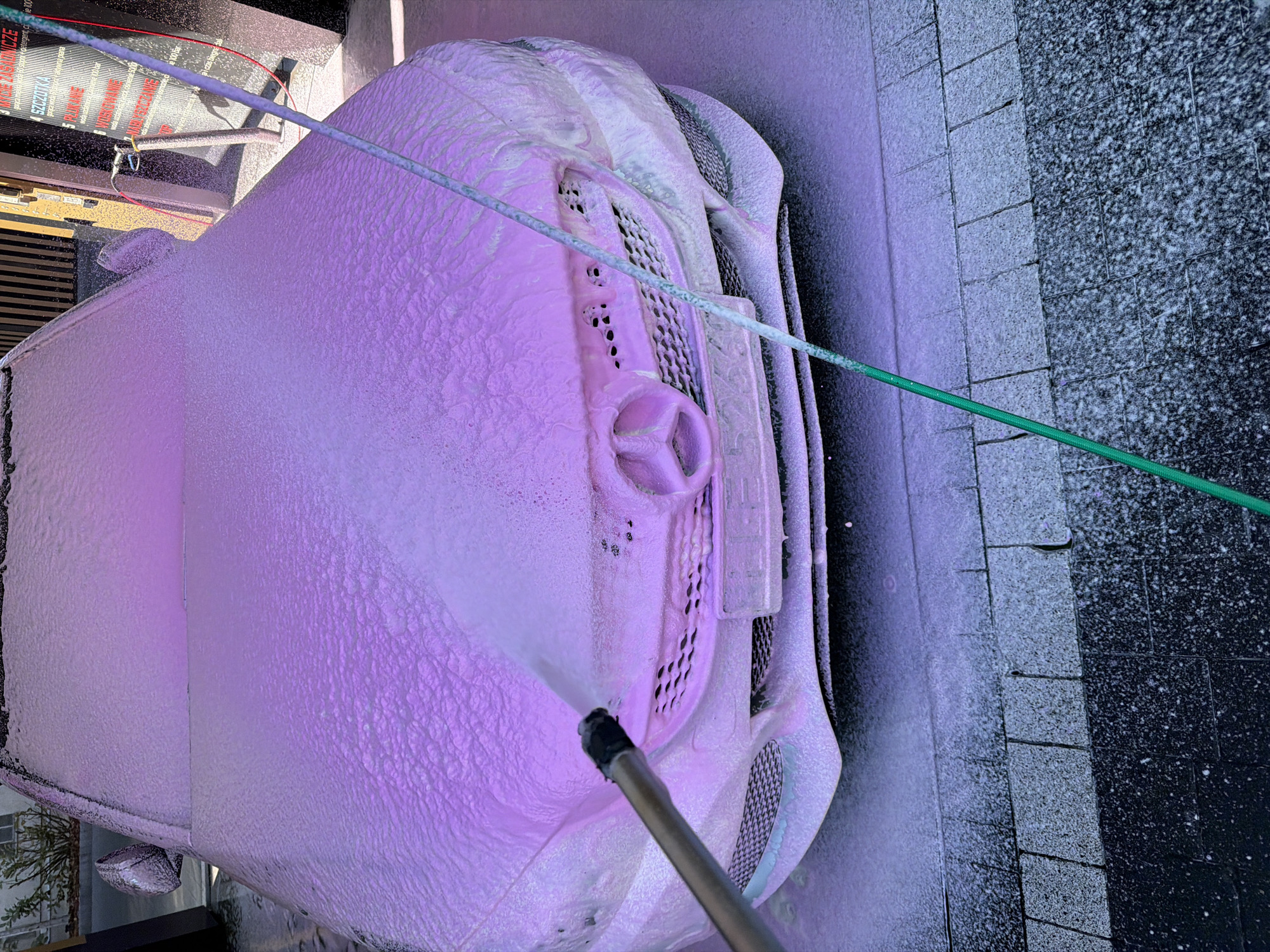 A top-down shot depicts a car entirely covered in pink soap foam, being washed by a person holding a pressure washer wand. The car, identified by its Mercedes-Benz logo on the grill, is parked on a paved surface, presumably at a car wash or a residential driveway. A green hose is visible extending away from the car. The environment appears urban, with building structures visible in the background. The location is unknown, as it is unspecified in the prompt.