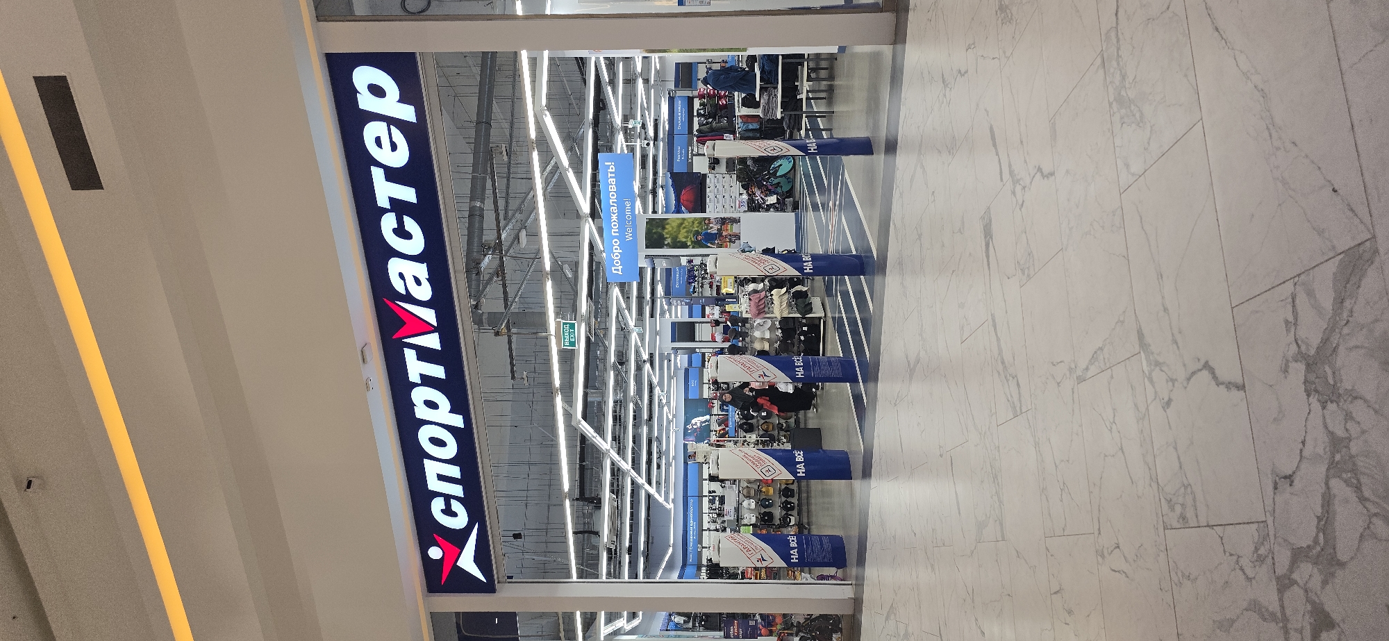 The image shows the entrance to a "Sportmaster" store, a sporting goods retailer. The store name is prominently displayed in Cyrillic letters above the entrance. The interior of the store is visible, with racks of merchandise and signage. There are blue pillars along the entrance with white text that says "HA BCE" which means "Everything is on sale". There is a sign above the store displays that says "Добро пожаловать! Welcome!" which is Russian and English for "Welcome!". The store is in a shopping mall. The flooring outside the store is white marble-like tile. There is a person wearing a black coat looking at goods on display. There is a sign that says "Exit" in Russian and English. The mall has fluorescent tube lighting. The exact location and time are unknown, but the image was taken at a store in the city of Grozny, Russia.
