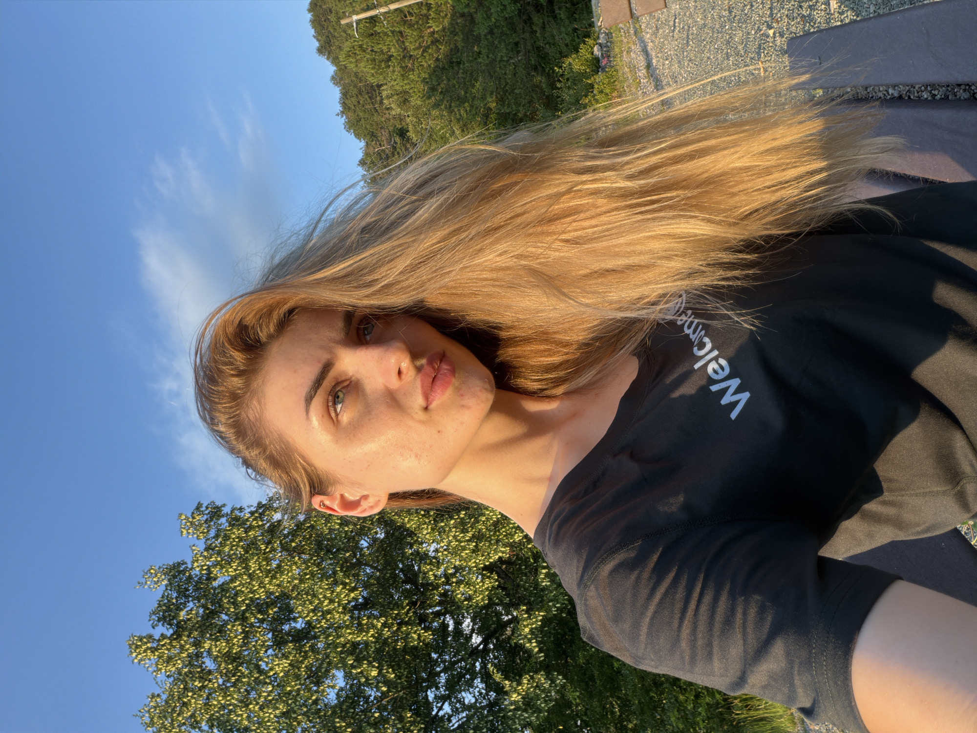 A selfie of a young woman with long, blonde hair and fair skin is taken outdoors. She is wearing a black t-shirt with the word "Welcome" in white lettering. The woman is looking slightly upwards and to the side, with her hair falling across her face and shoulder. She has freckles on her face and her ears are visible. The background features a bright blue sky with some wispy clouds, green trees, and a wooded hillside. The lighting suggests it is either early morning or late afternoon, with the sun casting a warm glow on the scene. The ground appears to be gravel.