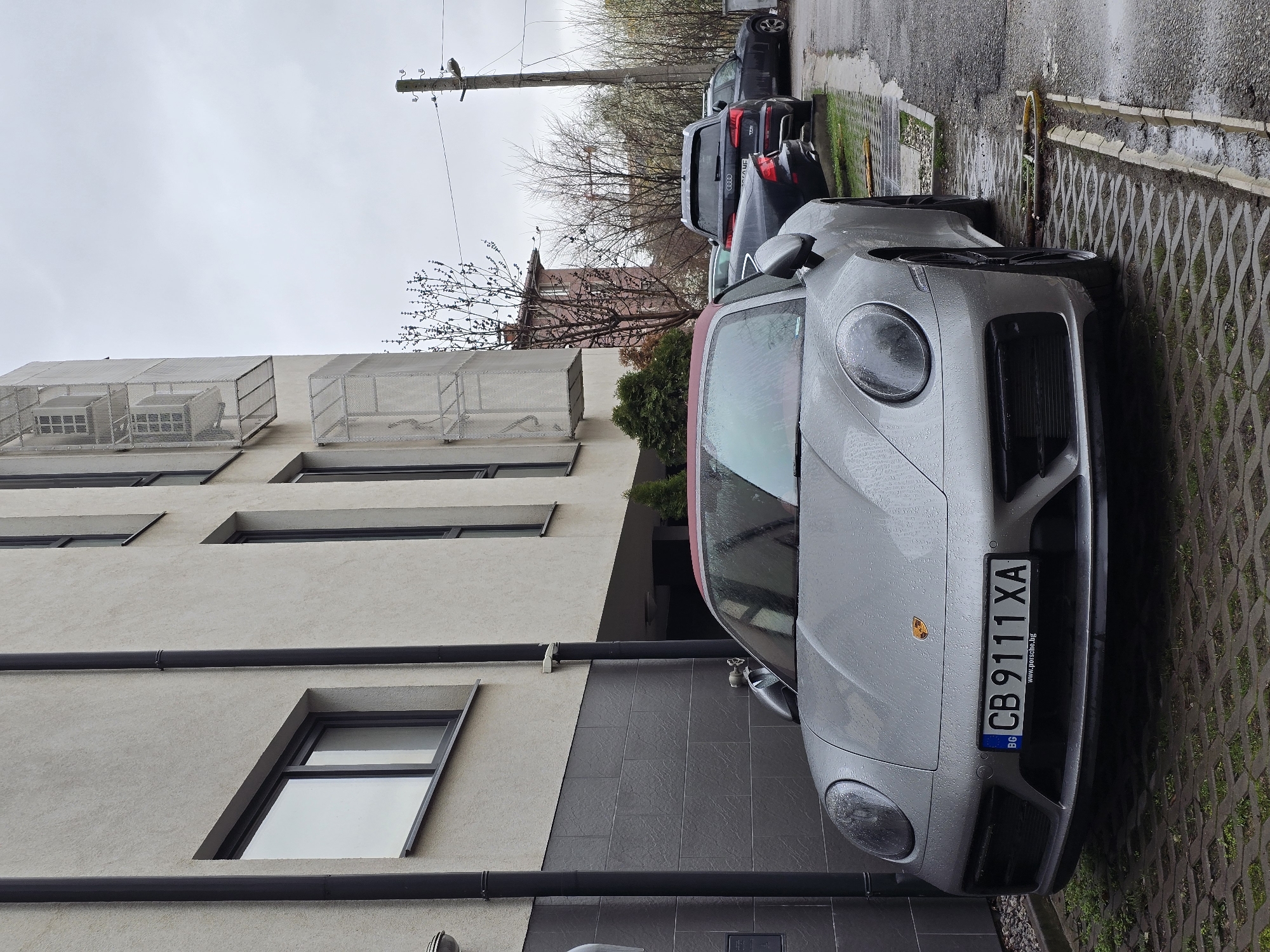 The image shows a silver Porsche parked in front of a modern building. The license plate of the car reads "CB 9111 XA," which is a Bulgarian license plate, suggesting the location is in Bulgaria. The building is multi-story with a light-colored facade, dark-framed windows, and metallic screened enclosures. The area seems to be a residential or mixed-use neighborhood. The ground next to the car is paved with a patterned concrete block. The weather appears overcast and wet, evidenced by water droplets on the car's surface. The presence of other parked cars further back suggests the scene is on a public street or parking area. The scene is in Sofia, Bulgaria.