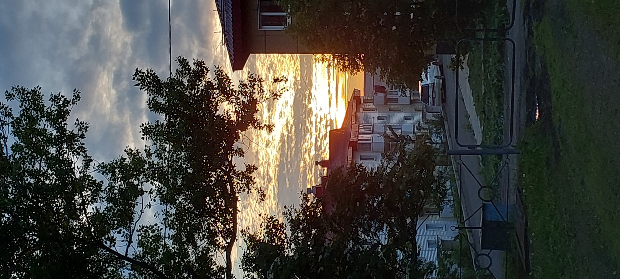 The image depicts a sunset scene in Aleksandrovsk-Sakhalinskiy, Russia. In the foreground, there is a dark grassy area with a metal fence. Behind the fence, a road curves, bordered by green grass and trees. Several multi-story residential buildings with white facades and red roofs are visible in the middle ground. A white van is parked on the road. The sky is filled with dramatic clouds illuminated by the setting sun, which casts a warm golden glow. The overall impression is that of a residential neighborhood at dusk.