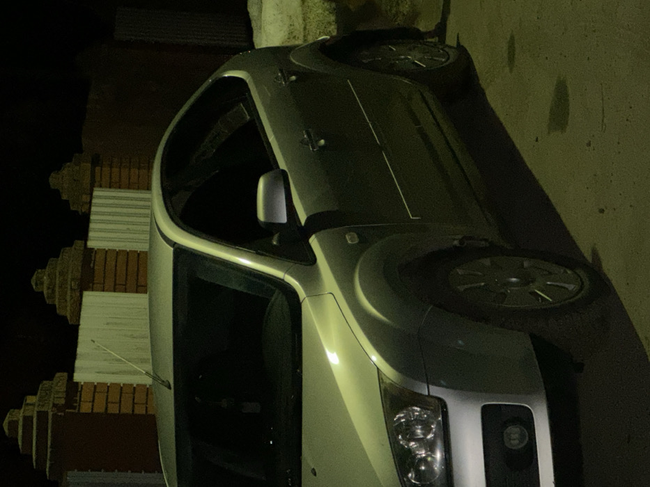 A silver Ford Focus hatchback is parked on a roadway during nighttime hours in Iksha, Russia. The vehicle, a second-generation model, is positioned with its front left quarter and entire left side visible, illuminated by an off-camera light source that casts a prominent yellowish-green hue onto the vehicle's upper surfaces and the asphalt ground, creating a distinct shadow. An antenna is present on the roof, and an air freshener or ornament hangs inside the front windshield. In the background, a fence composed of brick pillars with stepped caps and light-colored, possibly corrugated metal panels is visible. To the right, a darker wall structure and a patch of light-colored ground or snow are partially visible behind the vehicle. The ground exhibits minor irregularities and dark stains. No individuals or dynamic actions are depicted.
