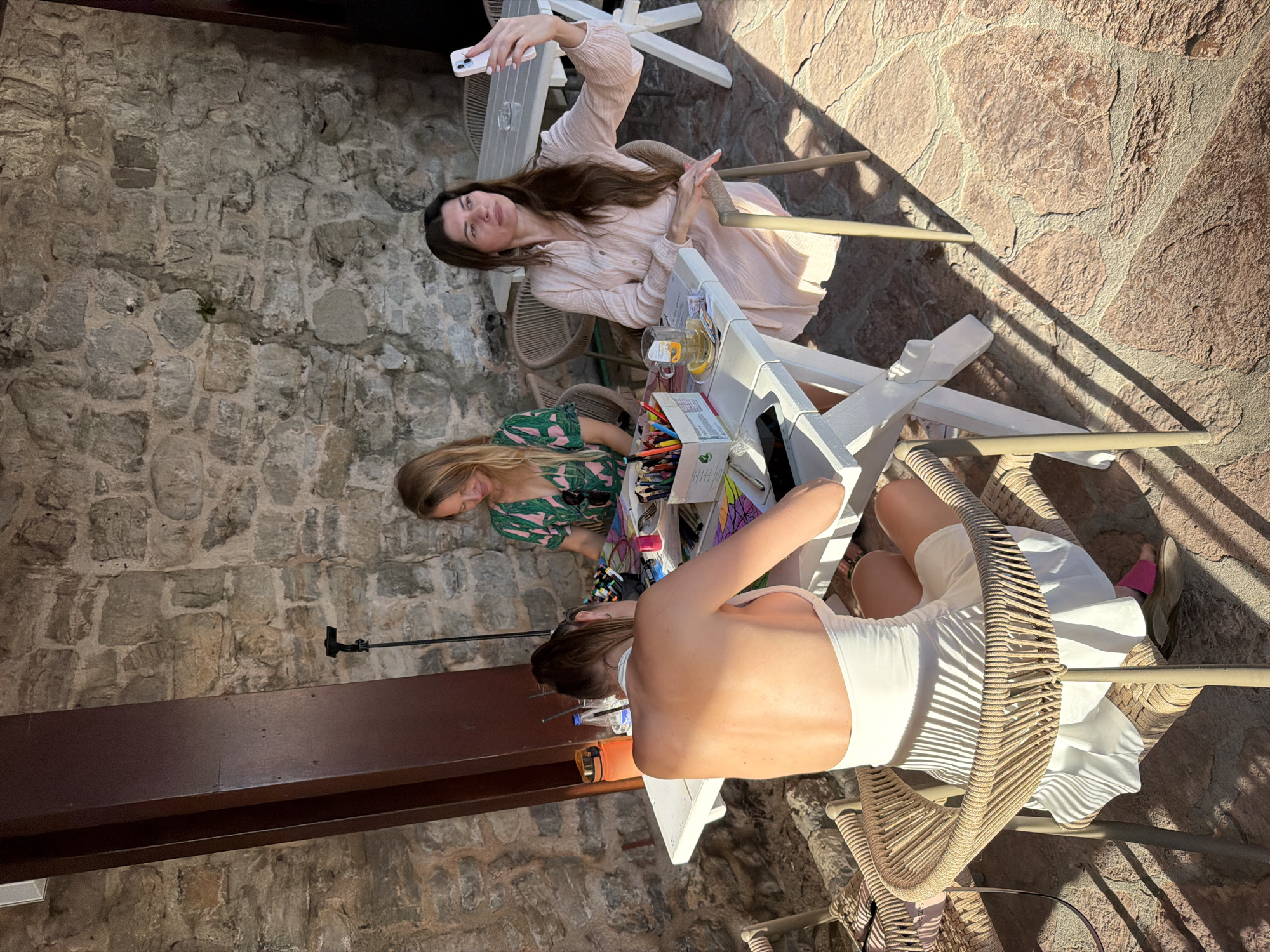 Three women sit around a white table in what appears to be an outdoor café or patio. The first woman, seated at the top of the frame, is holding a white smartphone up as though taking a selfie. She has long brown hair and is wearing a light pink long-sleeved dress. Below her, a second woman is seated at the table wearing a green and pink printed top. She is leaning over the table, seemingly focusing on something on the table surface. The third woman is seated at the bottom of the frame and is wearing a white dress or top, revealing her bare back. The table is covered with items, including what appears to be a box of colored pencils or markers, papers, beverages, and other miscellaneous objects. All three women are seated in wicker chairs. The area is walled with old stone and brick and the ground is paved with stone. Shadows fall across the patio, indicating bright sunlight. The location appears to be the Old Town of Budva, Montenegro, judging by the architecture and stone walls.