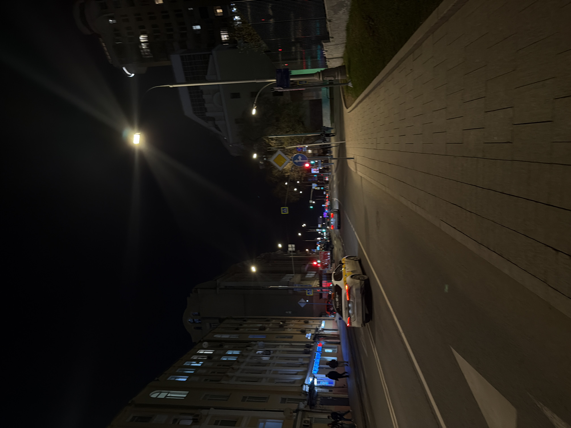 The image depicts a street scene at night in Moscow, Russia. The perspective is from the side of the road, looking down the street. The street is illuminated by streetlights and the lights of buildings. A car is stopped on the side of the street. Pedestrians are walking on the sidewalk. The buildings are tall and densely packed, typical of an urban environment. A row of low-rise buildings lines the street to the left. Signage and street signs are visible along the street. The road surface is asphalt. A brick paved sidewalk stretches off to the right. The sky is dark.