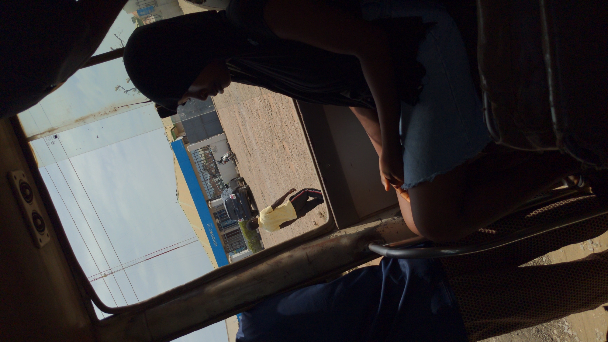 The image captures a perspective from inside a vehicle, looking out onto an urban street scene during the daytime. In the foreground, a person wearing a dark head covering and a dark top over denim shorts or a skirt is partially visible, leaning forward. Elements of the vehicle's interior, including a speaker on the left and parts of its frame and patterned upholstery, define the immediate surroundings.
Beyond the vehicle, a bright, clear sky fills the upper portion of the exterior view. Below the sky, several buildings line a street or pavement. A prominent building on the right features a distinct blue facade with white text that clearly spells out "Union Bank" and "unionbank.com." Parked vehicles, including a dark car and several motorcycles, are visible along the street. Another person, dressed in a yellow t-shirt and dark pants with red stripes, is standing on the pavement outside. The scene depicts a sunny day, showcasing a typical street environment, likely in Ado, Nigeria, given the visible bank branding.