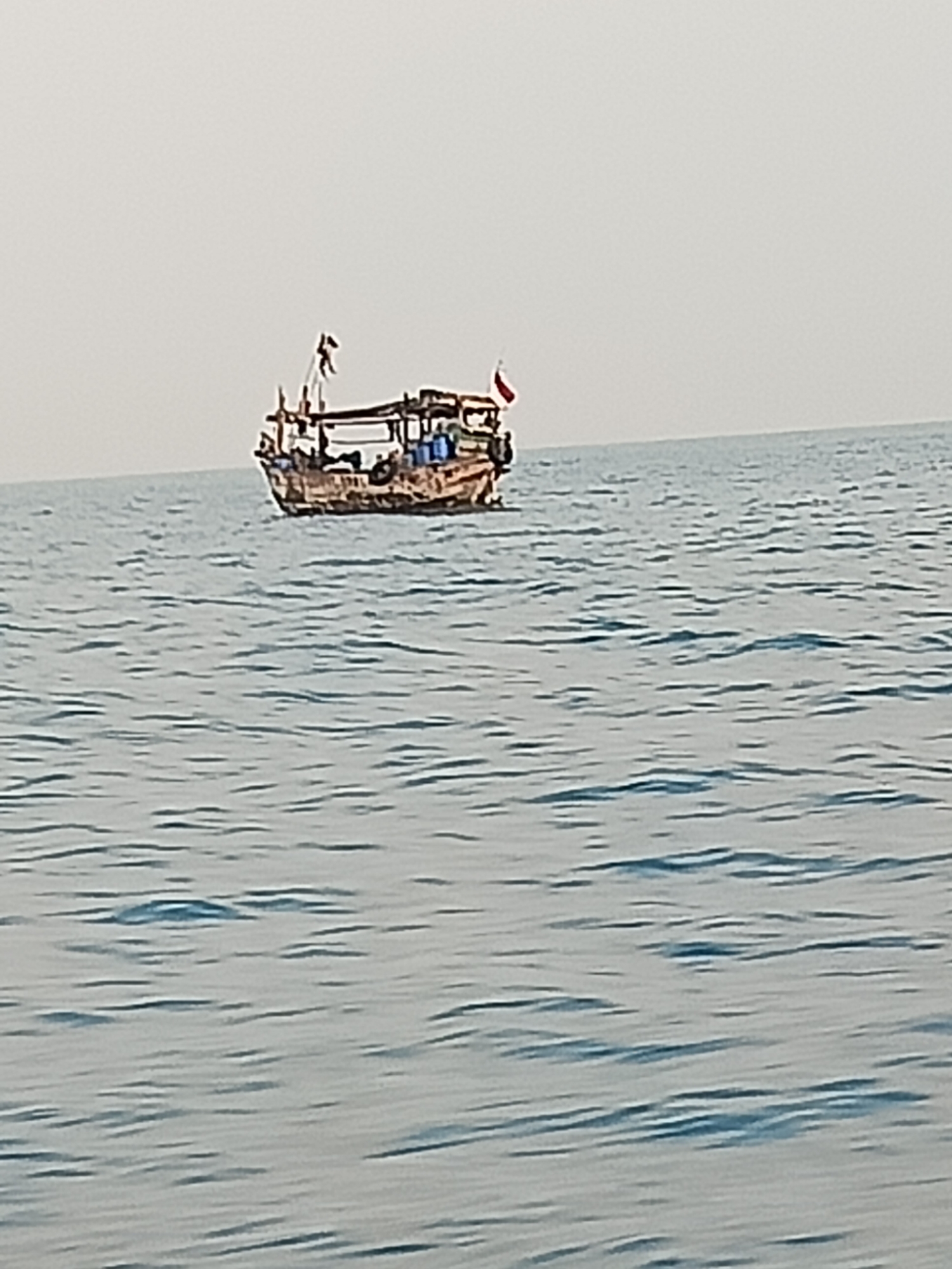 A small, weathered wooden boat is sailing on calm, blue water under a hazy sky. The boat has a thatched roof covering part of its deck and appears to be carrying some cargo or equipment. Two small flags, one red and one red with white markings, are visible on poles at the front of the boat. The boat is positioned in the middle ground of the image, with the water filling the foreground and a featureless, pale sky occupying the background.