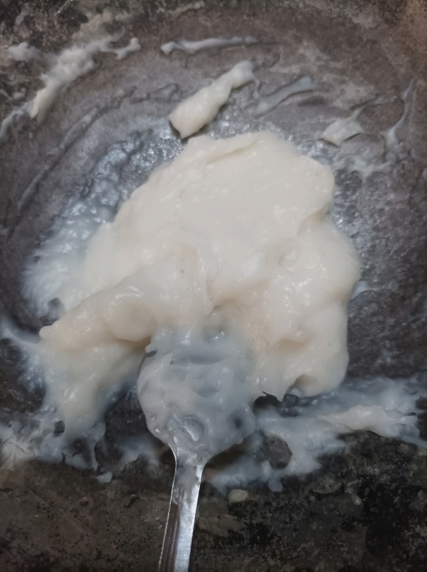 A close-up, overhead shot shows a metallic spoon partially submerged in a thick, creamy, off-white substance, likely a paste or batter. The substance is piled in the center of a dark, textured bowl or pan, with some of the substance spread thinly around the edges and adhering to the sides. The bowl's surface appears rough and slightly metallic, with variations in color from dark gray to lighter, almost silvery tones. Streaks and smears of the white substance are visible on the bowl's interior, suggesting it has been mixed or stirred. The lighting is overhead and appears to be artificial, creating subtle shadows and highlights on the creamy substance and the bowl's surface. The overall impression is of a domestic or culinary scene, likely indoors, with no indication of an outdoor environment or city location.