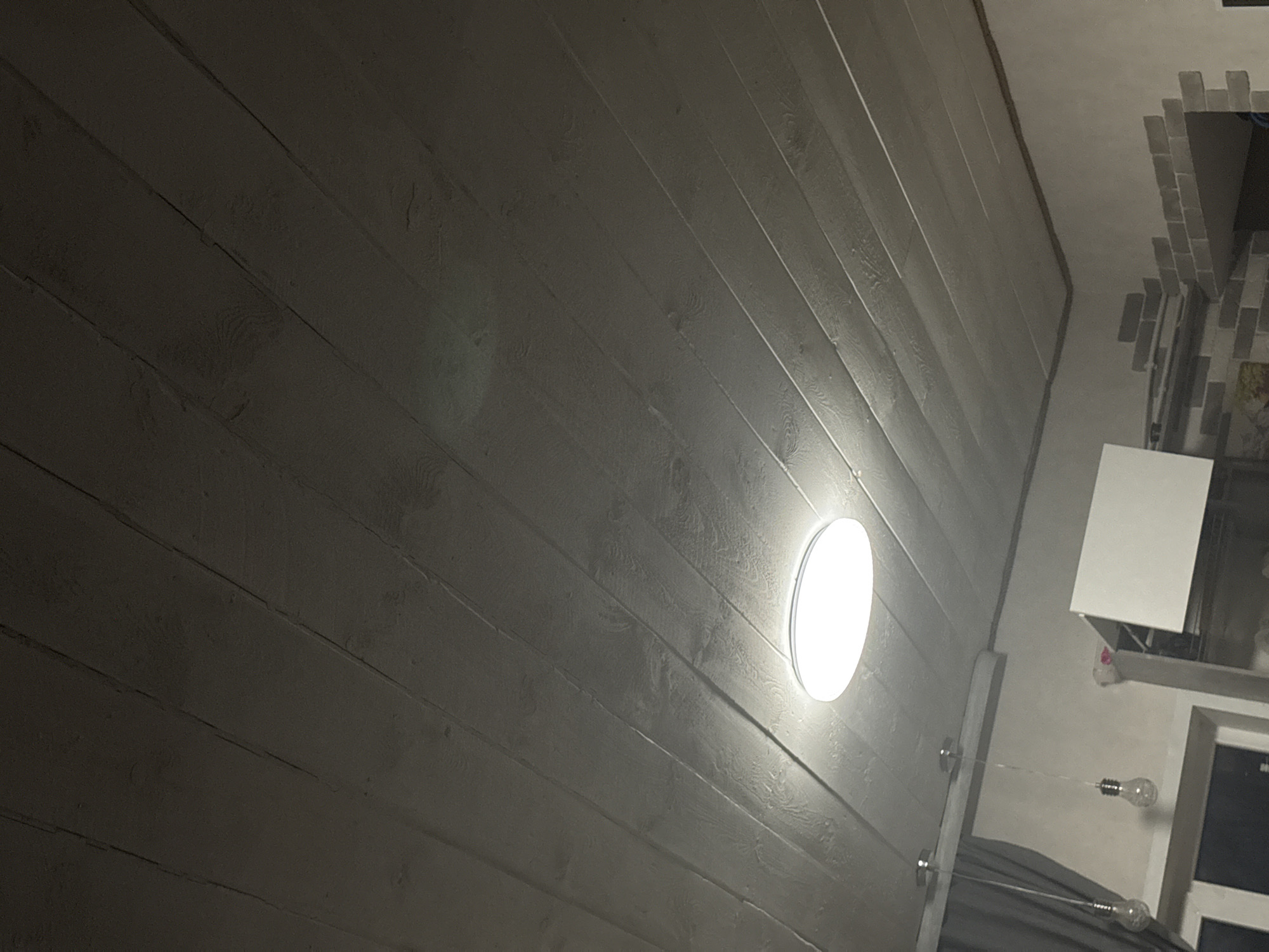 The image depicts an interior shot, focusing on a ceiling made of parallel wooden planks painted in a gray or off-white color. A round ceiling light fixture is illuminated, casting a bright glow. In the upper right corner, a section of decorative brickwork is visible, alongside a square, white object that appears to be part of the room's decor or architecture. Below this, a window with a dark interior is present. Additionally, there are two hanging light bulbs suspended from the ceiling by thin wires. The location is Chaikovsky, Russia.