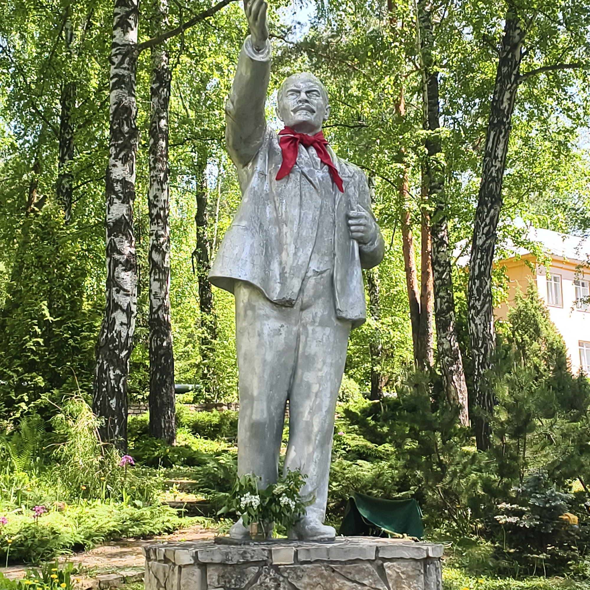 This image shows a statue of Vladimir Lenin located in the city of Варзи-Ятчи, Russia. The statue is made of a silver-colored material and depicts Lenin in a suit with a red scarf around his neck, his right arm raised. The statue stands on a stone base and is surrounded by trees and greenery. A yellow building can be seen in the background.