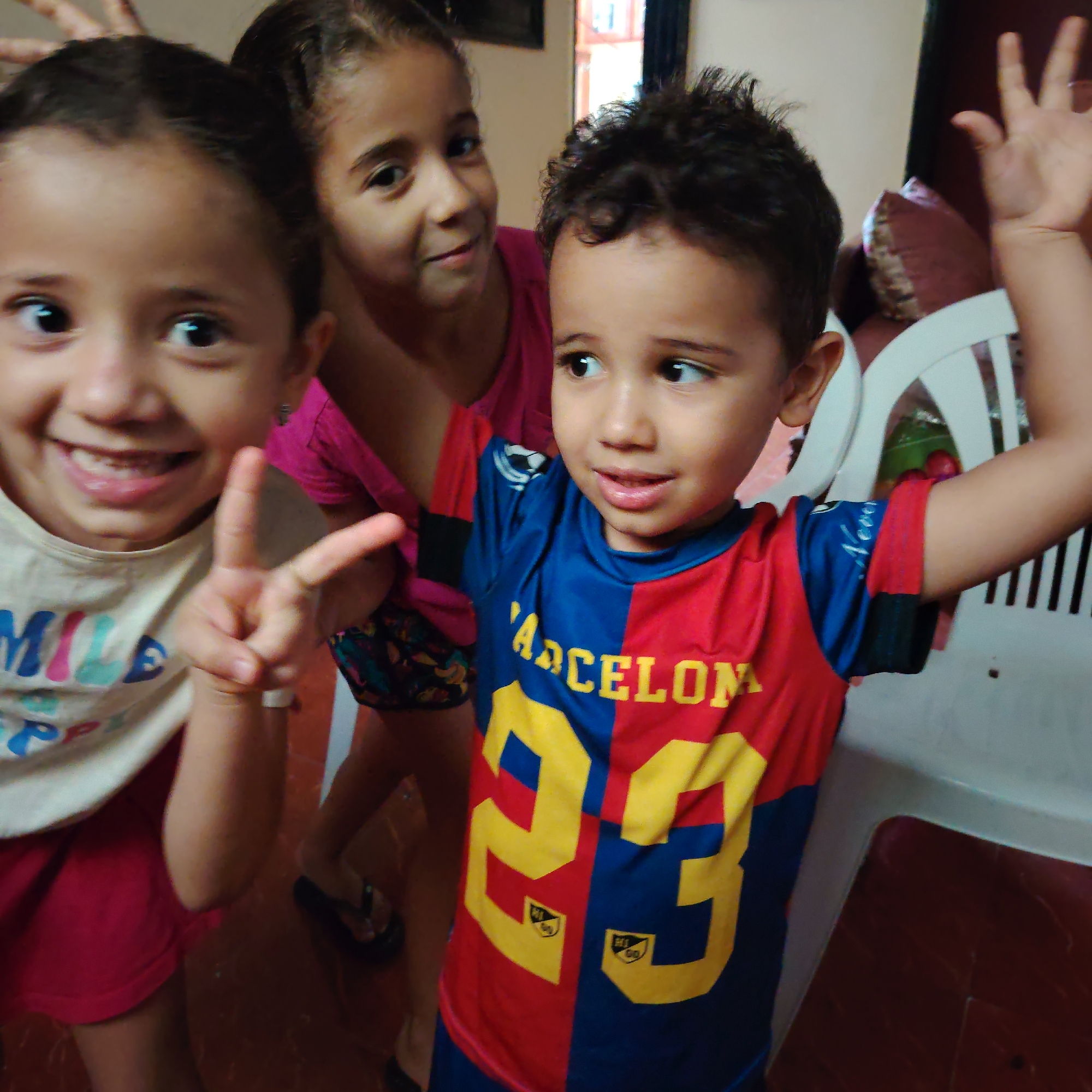 In Ouaouizert, Morocco, a group of three young children are captured in a candid moment indoors. The child on the left, facing the camera, wears a white t-shirt with colorful lettering and red shorts. They are smiling broadly and making a peace sign with their right hand. To their right, a second child, wearing a pink t-shirt, is partially visible, looking towards the camera with a subtle smile. The third child, in the foreground on the right, is wearing a red and blue soccer jersey with the number "23" prominently displayed. This child is looking off to the side, with one hand raised in a wave. The background is indistinct, but appears to be an indoor setting with hints of furniture and wall decor. The lighting suggests an interior space with ambient light.