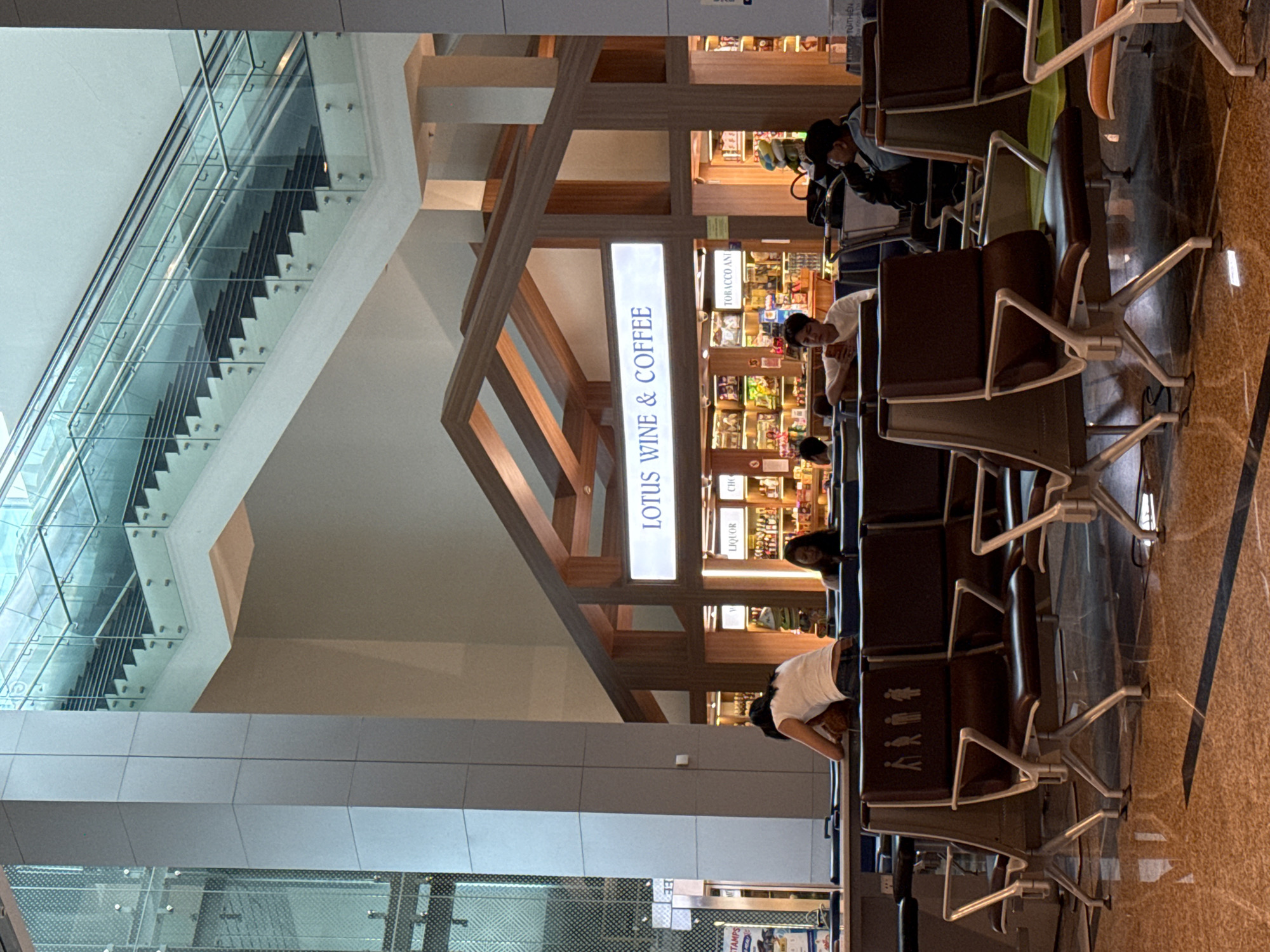 The image depicts an interior scene, likely an airport terminal in Bắc Cam Ranh, Vietnam, given the presence of seating areas and a duty-free shop. In the foreground, rows of dark brown airport-style seating with metallic legs are visible, some featuring a stencil of four figures, two male and two female, on the backrests, possibly indicating unisex restrooms. The floor is a polished, speckled brown material reflecting the ambient light.
Further back, a structure with a wooden, A-frame roof houses a brightly lit shop. A sign above the shop reads "LOTUS WINE & COFFEE". The shop's interior displays shelves stocked with various products, including bottles of liquor. Various individuals are seated or standing within the scene. One person, wearing a white top and dark pants, is leaning over a table in the seating area, facing away from the camera. Another person in a light-colored shirt is seated in the mid-ground, looking at a mobile phone. Others are seated further back, some appearing to be engaged in conversation or waiting.
In the background, a modern multi-story structure with glass railings and escalators is visible, suggesting multiple levels within the terminal. The overall lighting is artificial and bright, casting shadows on the floor.