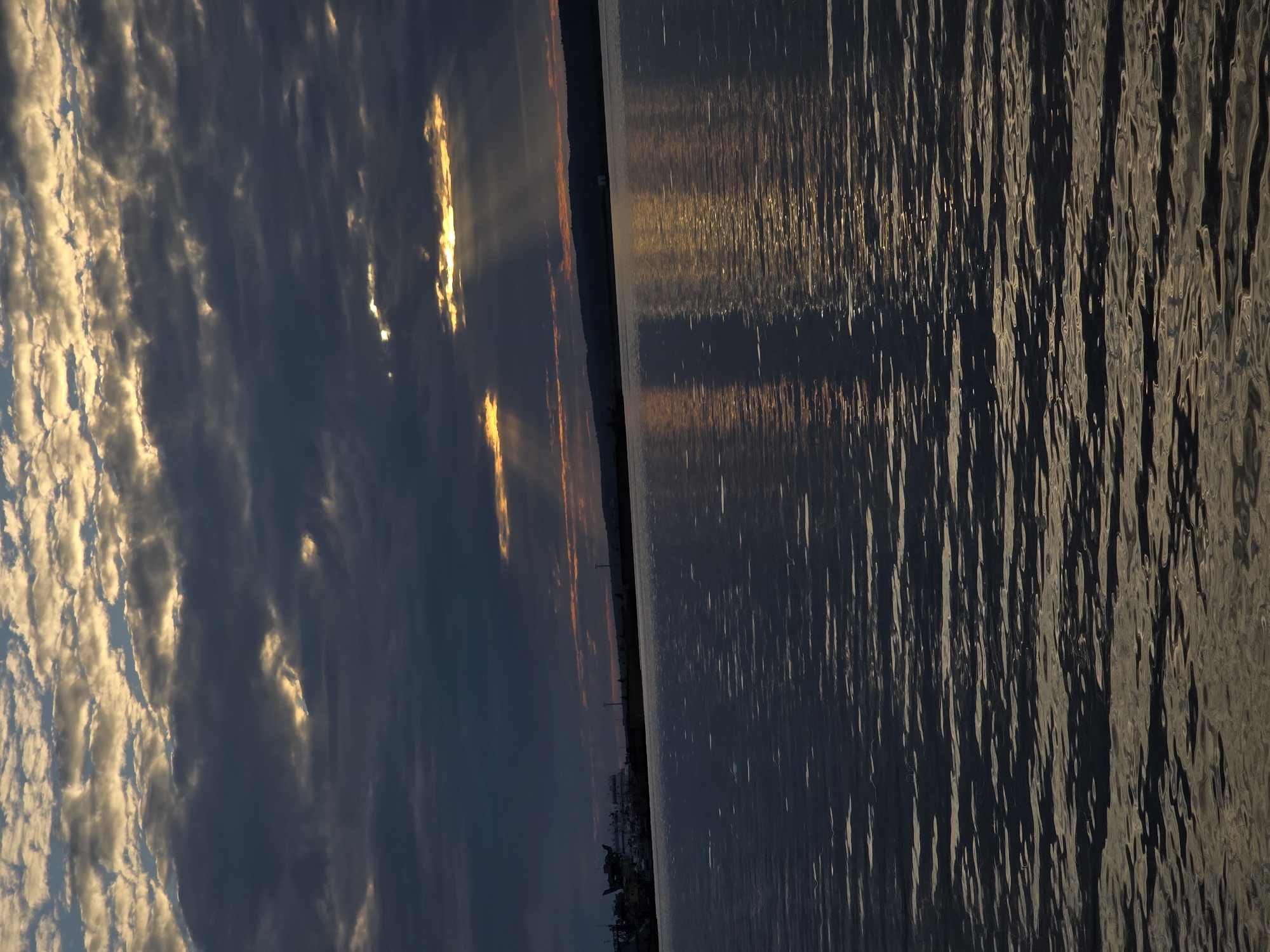 The image captures a body of water, possibly a river or reservoir, with a shoreline visible in the distance. The sky is filled with dramatic clouds, with shafts of sunlight breaking through, illuminating the water's surface with golden reflections. On the left side of the distant shoreline, several ships or boats are docked at a port, with structures and buildings visible behind them. The overall impression is one of natural beauty and industrial activity coexisting. The location is identified as Chaikovsky, Russia.
