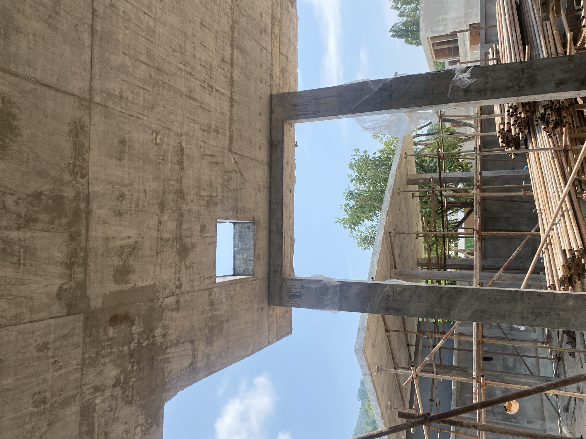 The image depicts the unfinished concrete structure of a building under construction, likely in Choeng Thale, Thailand, as suggested by the environment and architectural style. The view is angled upwards, showcasing the raw concrete walls, beams, and scaffolding. A rectangular opening serves as a window on the left wall. The right side shows scaffolding made of metal pipes surrounding the building, with stacked pipes adjacent to the structure. Outside the building, green trees are visible through the scaffolding, and the sky appears blue with scattered clouds. There are no people visible in the frame. The overall environment is that of an active construction site.