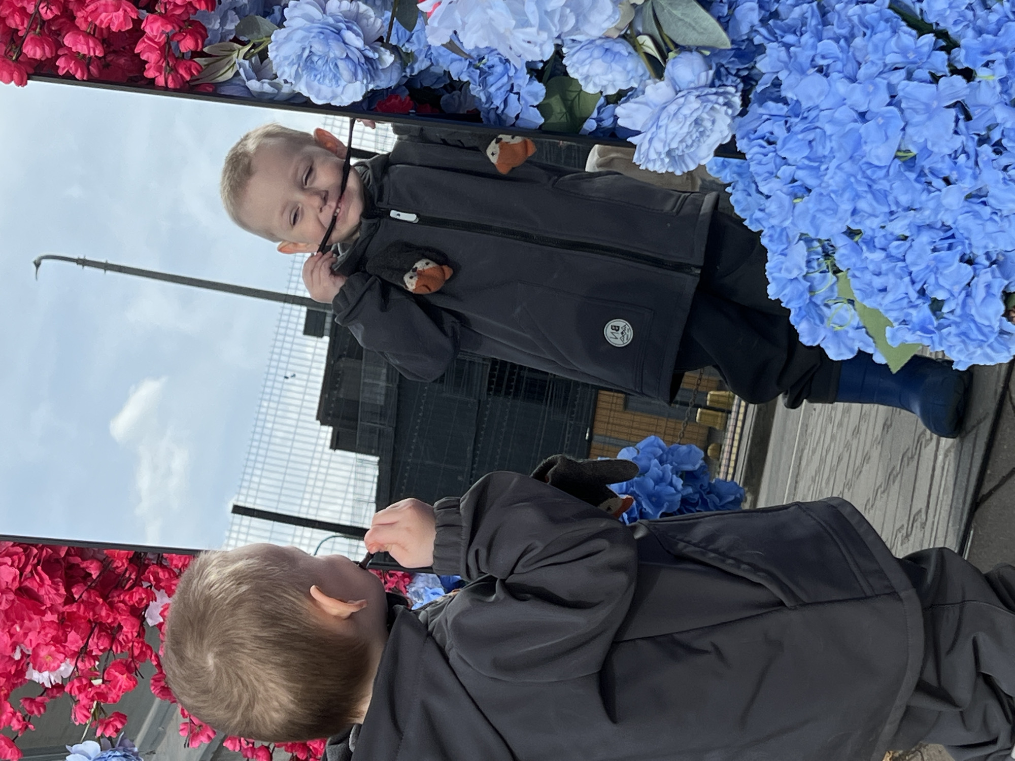 A young child in a black jacket and blue boots stands in front of a mirror decorated with pink and blue artificial flowers. The child is reflected in the mirror, and the reflection shows a building in the background. The photograph was taken in Mytishchi, Russia, based on visual cues like the building in the reflection. The child is holding something in their mouth, possibly a string or a small object.