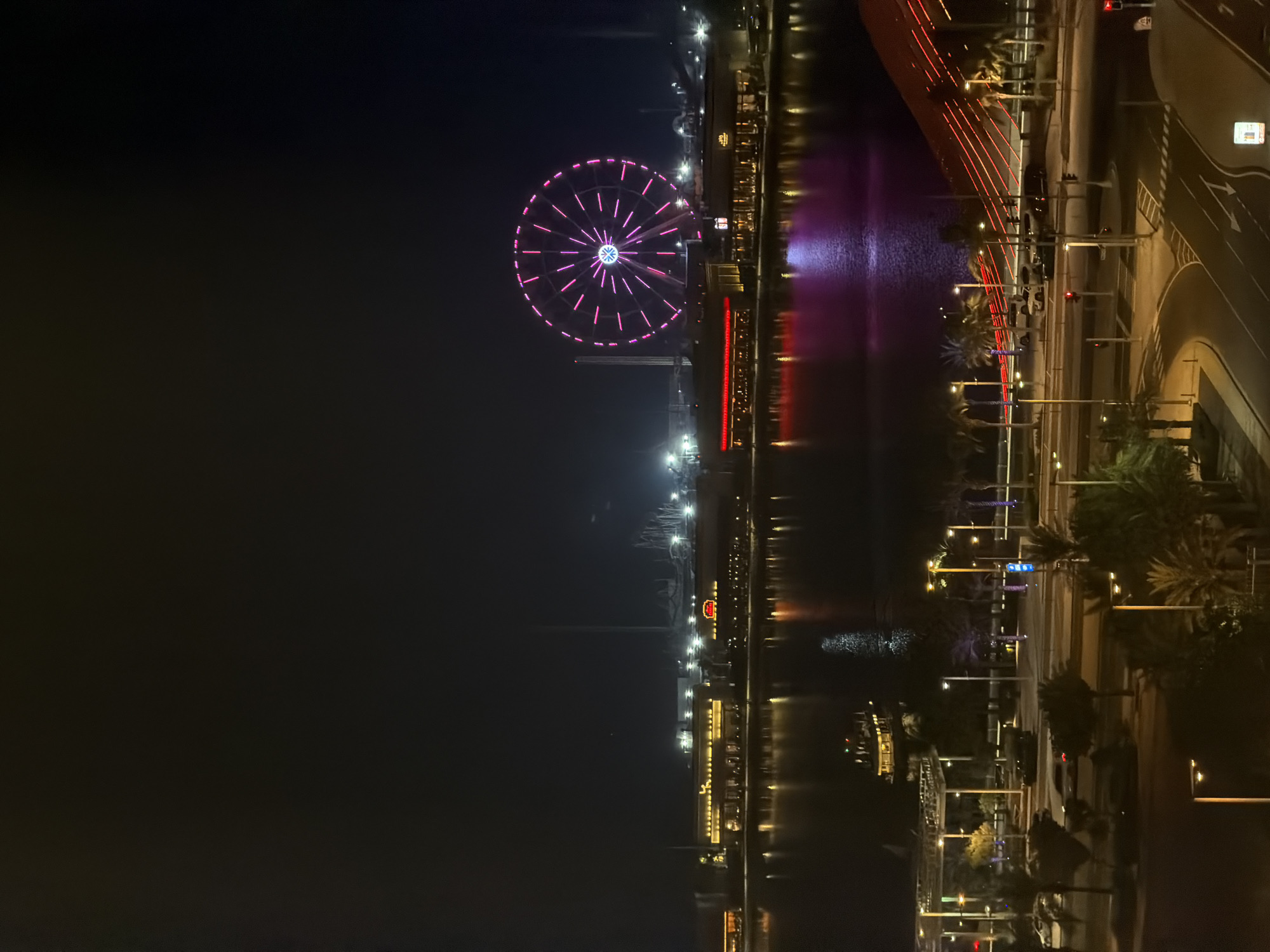 An elevated nighttime view captures a waterfront city scene illuminated by artificial lights. A large Ferris wheel, brightly lit with pink and purple LEDs, dominates the upper right quadrant of the image, its spokes radiating outward. Below it, a row of buildings with glowing windows and red accents lines the waterfront. The water reflects the colorful lights of the buildings and the Ferris wheel, creating a shimmering spectacle. On the left side of the frame, a boat with illuminated decks navigates the water. In the foreground, a street with palm trees and streetlights is visible. Cars are present on the street, and some buildings and infrastructure are subtly lit. The overall impression is of a vibrant urban environment at night, with a focus on entertainment and illuminated architecture.