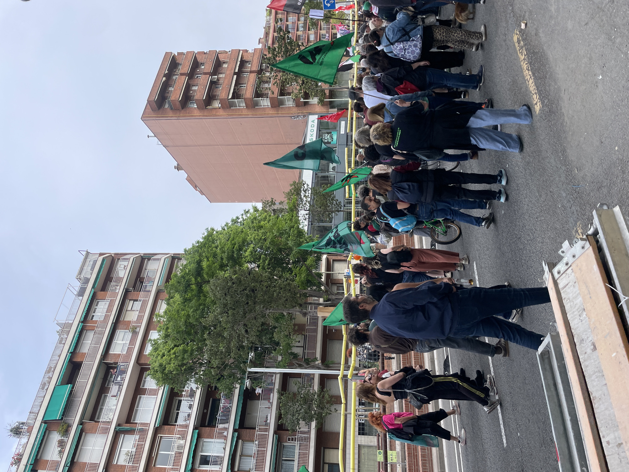 A crowd of people is gathered on a city street, seemingly participating in a demonstration or march. Many people are holding green flags, and the flags appear to have some type of emblem or insignia on them. The group is walking towards the right in the image. Buildings flank both sides of the street; one structure is a tall, light brown apartment or office building with recessed balconies. On the other side of the street is a light-colored building with dark brown accents. The sky is overcast. Based on the style of architecture, the buildings, and other details, the location could be the city of Barcelona, Spain.