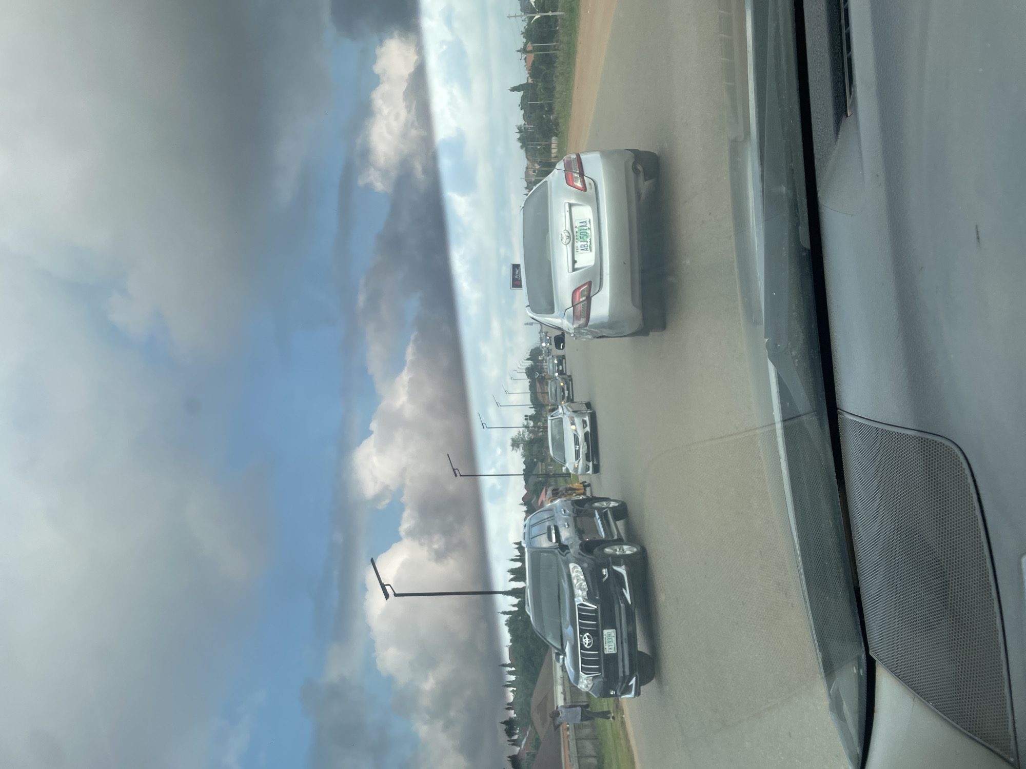The image shows a view from inside a vehicle looking out at a road with several cars. A black Toyota Land Cruiser Prado is in the left lane, with a license plate reading "EXY 933KL". To its right, a silver sedan is in the right lane with the license plate "ABJ 501AA". Further down the road, other vehicles are visible, as are streetlights and buildings on the sides of the road. The sky is partly cloudy. The scene appears to be in Galadimawa Village, Nigeria.
