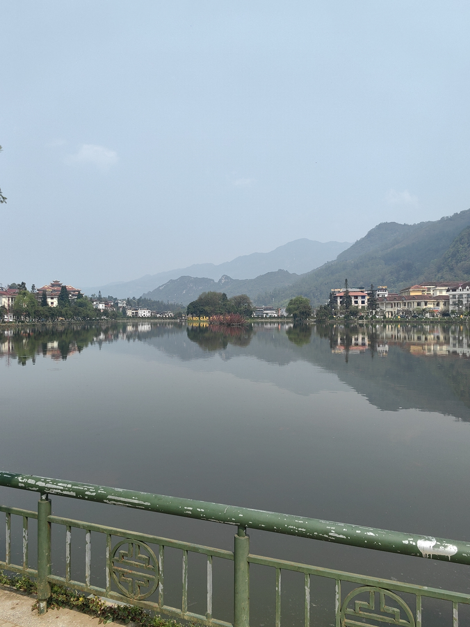 The image displays a panoramic view of Sa Pa Lake (Hồ Sa Pa) in Sa Pa, Vietnam, on a hazy day. In the foreground, a weathered green metal railing with peeling white paint extends across the bottom of the frame. The railing features circular decorative panels containing a stylized longevity symbol. Below the railing, a narrow strip of concrete ground with small weeds is visible.
The midground is dominated by the calm, dark surface of Sa Pa Lake, which reflects the surrounding structures and mountains. Along the lake's perimeter, a line of multi-story buildings with varied architectural styles—some featuring traditional tiered roofs and others a more contemporary design—are visible. Trees and dense foliage are interspersed among these buildings. An island or peninsula towards the center-right of the lake features thick vegetation and a large, illuminated yellow sign displaying the text "HANOI GÁP SA PA". Red-leaved shrubs are also present on this landmass.
In the background, successive layers of green mountains rise, receding into a light blue-grey atmospheric haze. The sky above is a uniform, pale blue-grey with minimal cloud definition. No individuals or active subjects are discernible in the scene.