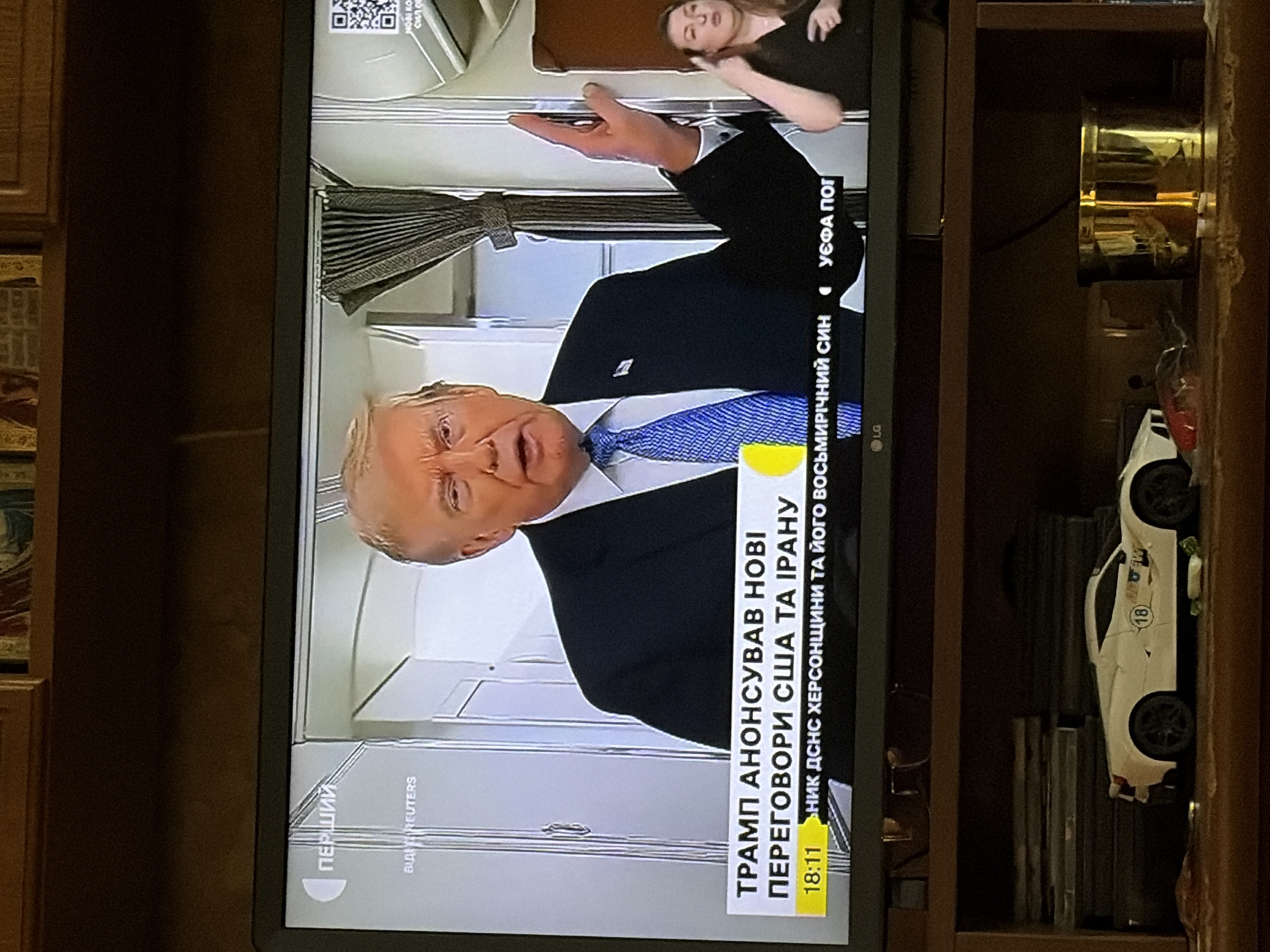 The image is a photograph of a television screen showing a news broadcast. On the screen, former U.S. President Donald Trump is speaking. He is wearing a dark suit and a blue tie. A news ticker at the bottom of the screen displays text in Ukrainian, which translates to "TRUMP ANNOUNCED NEW US-IRAN NEGOTIATIONS." To the right of Trump, a woman is visible, seemingly interpreting the broadcast in sign language. The background on the screen appears to be an interior setting. Below the television, on a shelf, a white toy sports car and a gold-colored object, possibly a mug or container, are visible. The environment appears to be a room in a home, with wooden furniture and shelves. The overall scene is indoors.
