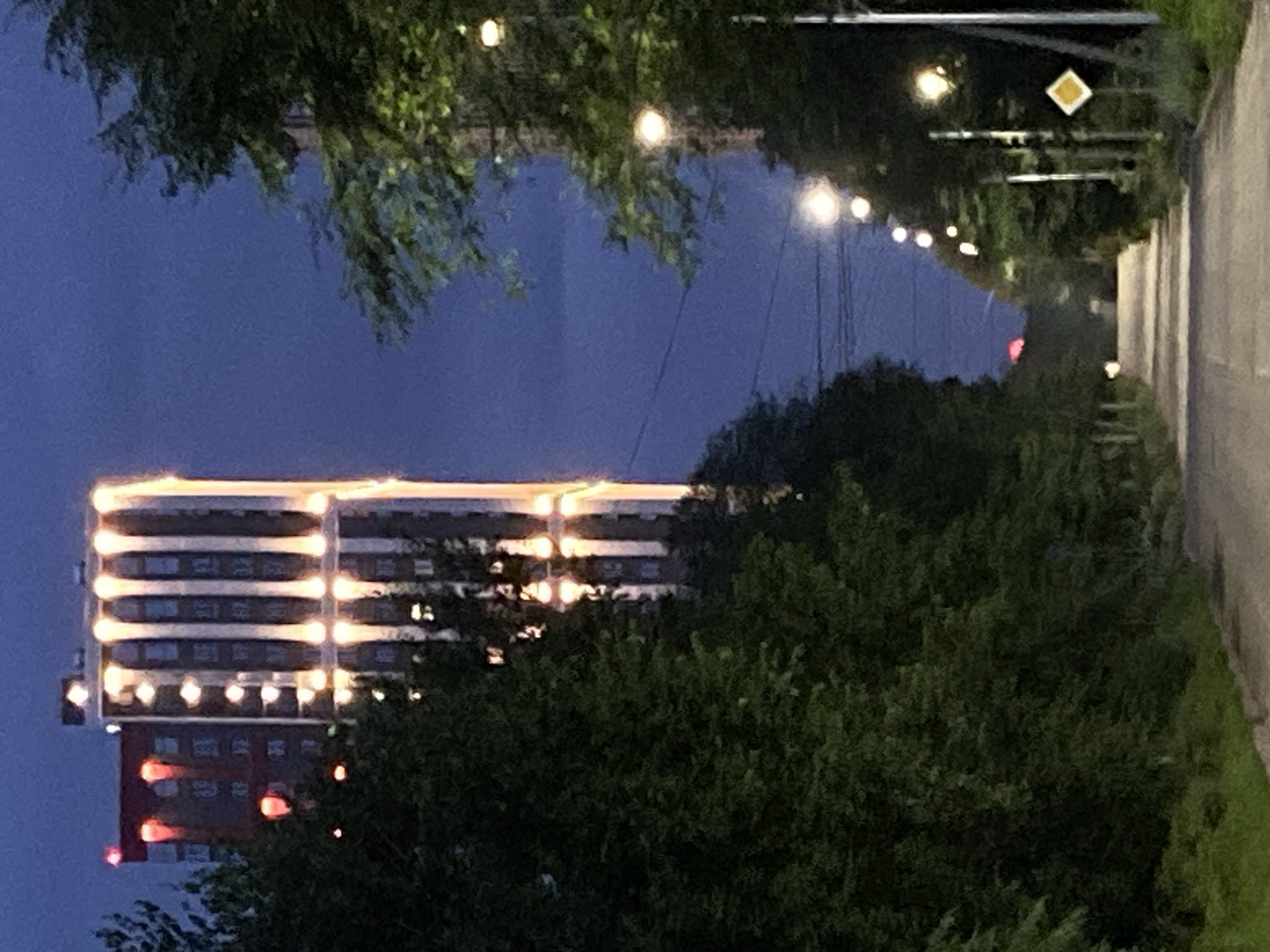 The image shows a street at dusk in Blagoveshchensk, Russia. On the left side of the street, there is a tall building with illuminated vertical sections. To the right of the building, trees and a street light are visible. The street itself is paved and curves towards the background. More trees line both sides of the street, with street lights illuminating the scene intermittently. A diamond-shaped yellow sign with a white border is affixed to a pole on the right side of the street. In the distance, a red light is visible, possibly from another building or a warning signal. The sky is a deep blue, characteristic of twilight.