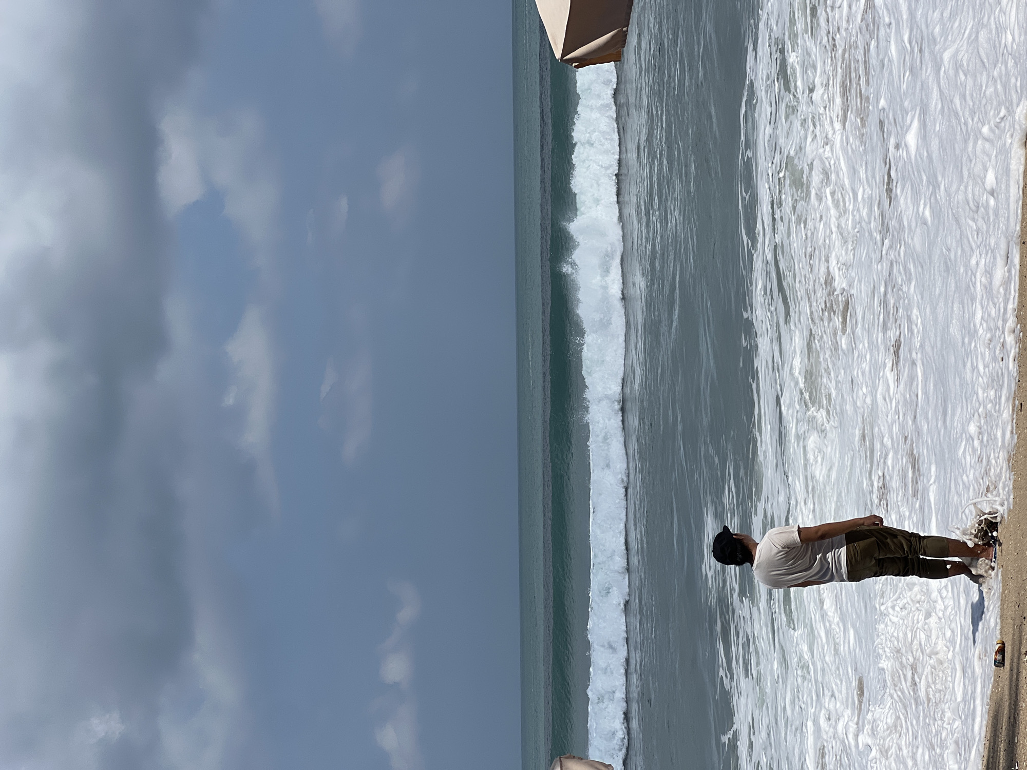A lone figure stands on the shore of Kuta Beach, Indonesia, facing a churning ocean under a partly cloudy sky. The individual, wearing a light-colored t-shirt and dark pants, is partially submerged in the foamy surf. The sea displays distinct layers of turquoise and darker blues, with a prominent white wave cresting in the middle ground. The beach itself is sandy, with a scattering of debris near the water's edge. A portion of a beige beach umbrella is visible on the right side of the frame, suggesting a resort or public beach setting. The overall atmosphere is one of contemplation amidst the powerful forces of nature.