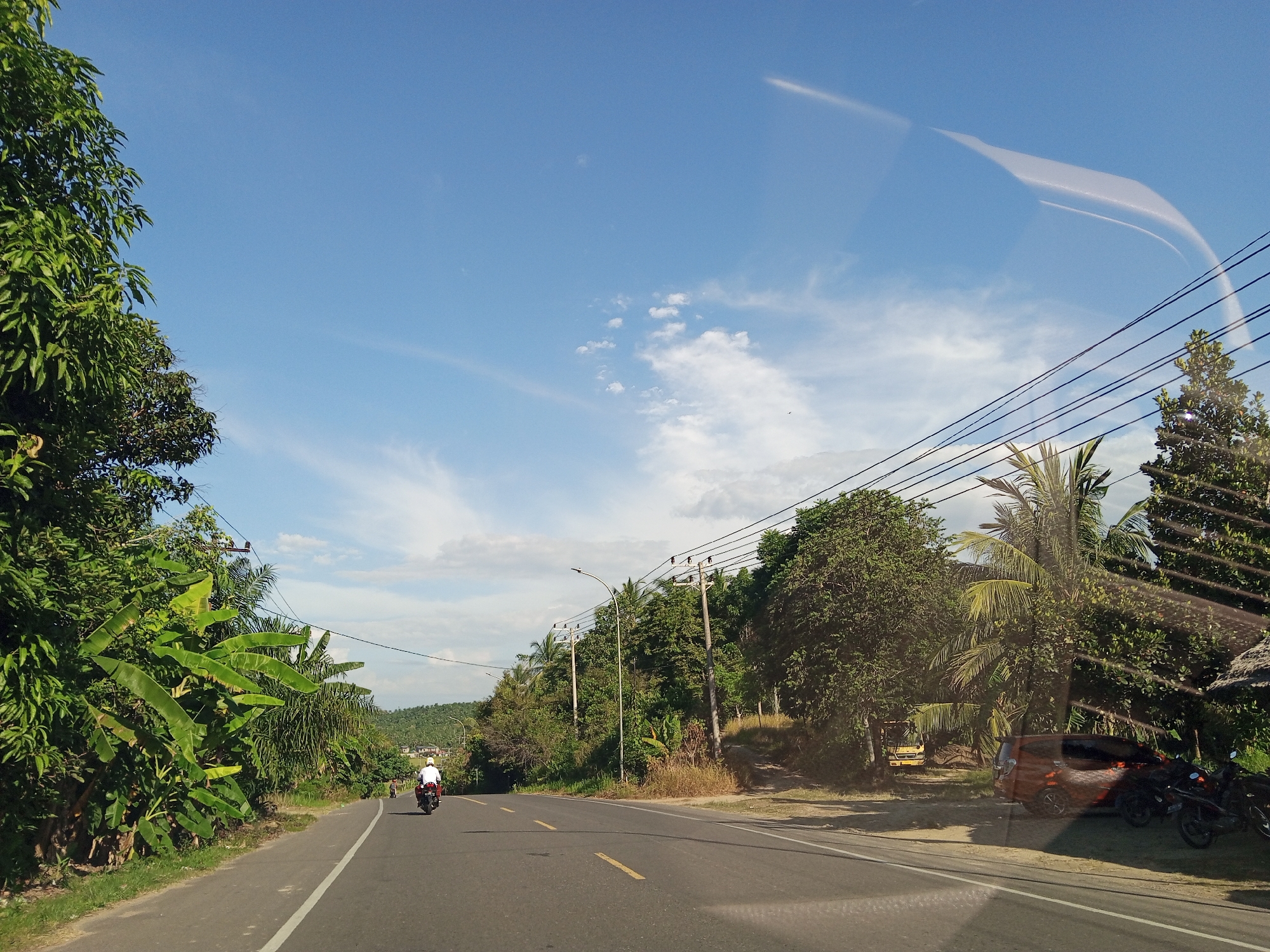 The image shows a road in Blangkarieng, Indonesia, during daylight. A person on a motorcycle is traveling away from the viewer on the asphalt road, which has a yellow dividing line. The road is lined with dense vegetation on both sides, including banana trees and other foliage. Utility poles and wires run along the right side of the road. A vehicle and motorcycles are parked on the right shoulder. The sky is blue with scattered clouds. There is a reflection on the camera lens.