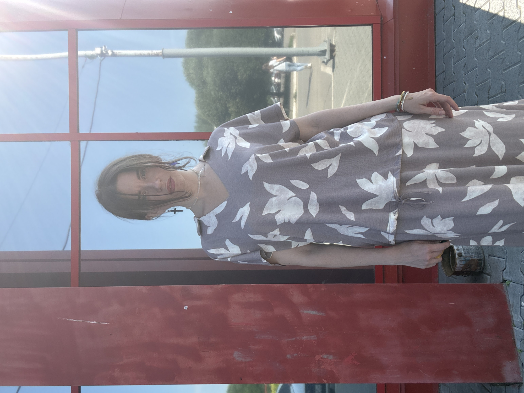 The image shows a woman with medium-length brown hair, wearing a grey dress with a white floral pattern. She is standing outside, reflected in a large window. The reflection shows a bright, sunny sky, suggesting it's daytime. The woman is looking towards the camera with a neutral expression. She is wearing a necklace with a cross pendant and several bracelets on her right wrist. Her left arm is extended, and her hand is near a metal object that appears to be a can or a pipe.
The background of the reflection shows trees and what looks like a parking lot with a few people in the distance. The foreground of the image features a red, metallic structure that frames the woman and her reflection. The ground is paved with grey tiles. The overall scene appears to be an urban or semi-urban outdoor setting. There is a slight glare from the sun on the window, which creates lens flare effects. The lighting suggests it is a bright, sunny day. There is no visible text in the image.