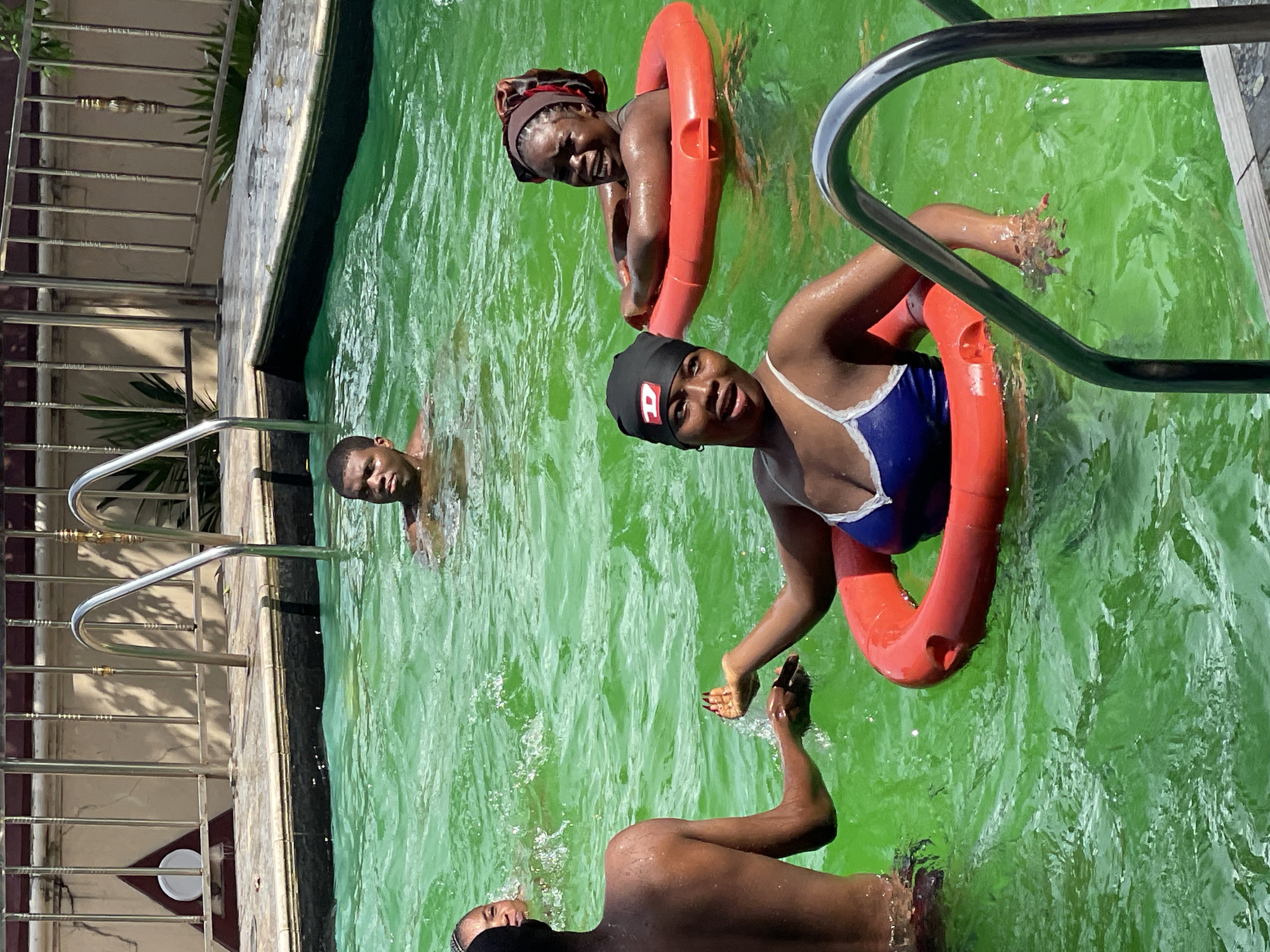 The image depicts a group of people enjoying a swimming pool. The water is a bright green color. In the foreground, a woman wearing a blue and white swimsuit and a black swim cap with a red logo is positioned in the center, with a red inflatable ring around her. She appears to be interacting with another person, whose back is to the camera and who is reaching out a hand towards her. To the right of the woman in the foreground, another woman, wearing a headscarf, is also in the pool, seemingly looking towards the person in front of her. In the background, a man is visible swimming in the pool, his head and shoulders above the water. The poolside has metallic railings and a tiled surface. The scene is outdoors, with sunlight casting shadows, indicating a daytime setting. The location is specified as Unknown, Unknown.