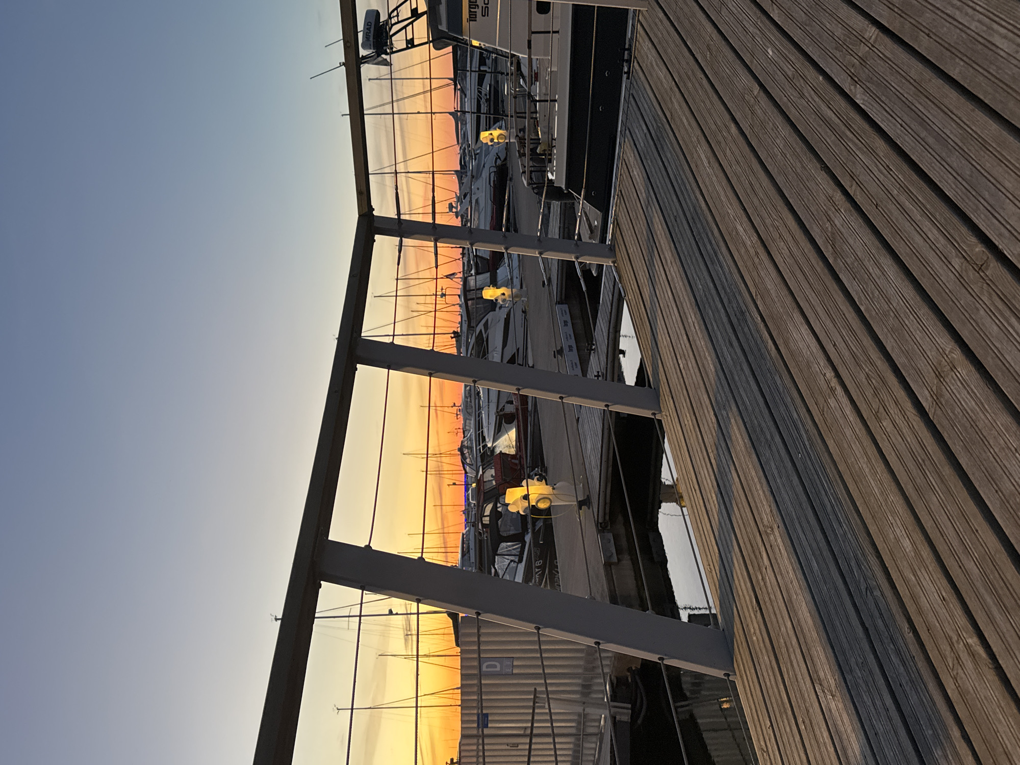 The image shows a marina at sunset, likely in Tallinn, Estonia, given the context. The foreground is dominated by a wooden deck, bathed in the warm light of the setting sun, casting long shadows. Beyond the deck, a metal railing with diagonal supports separates the viewer from the marina. The marina itself is filled with numerous boats, including sailboats with tall masts and motorboats. Some of the boats have their lights on, emitting a warm yellow glow. In the background, the sky transitions from a deep orange near the horizon to a clear blue overhead, characteristic of a sunset. The water in the marina reflects the sky and the lights of the boats. The overall scene conveys a sense of tranquility and the end of a day.
