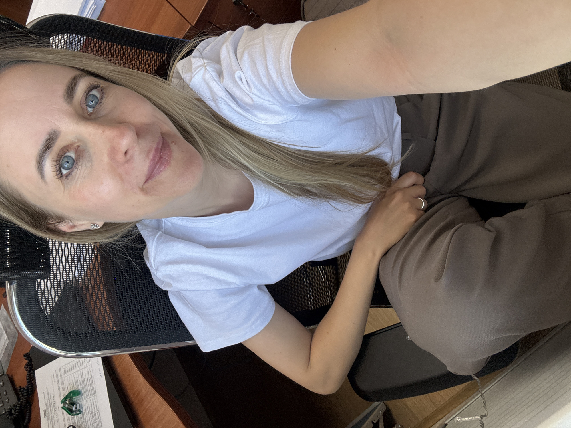 A woman with fair skin and blue eyes sits in a black office chair. She wears a white t-shirt and brown pants. She appears to be indoors, seated at a wooden desk. Papers, a stapler, and a telephone are visible on the desk. The location is potentially Chaikovsky, Russia, based on request context.