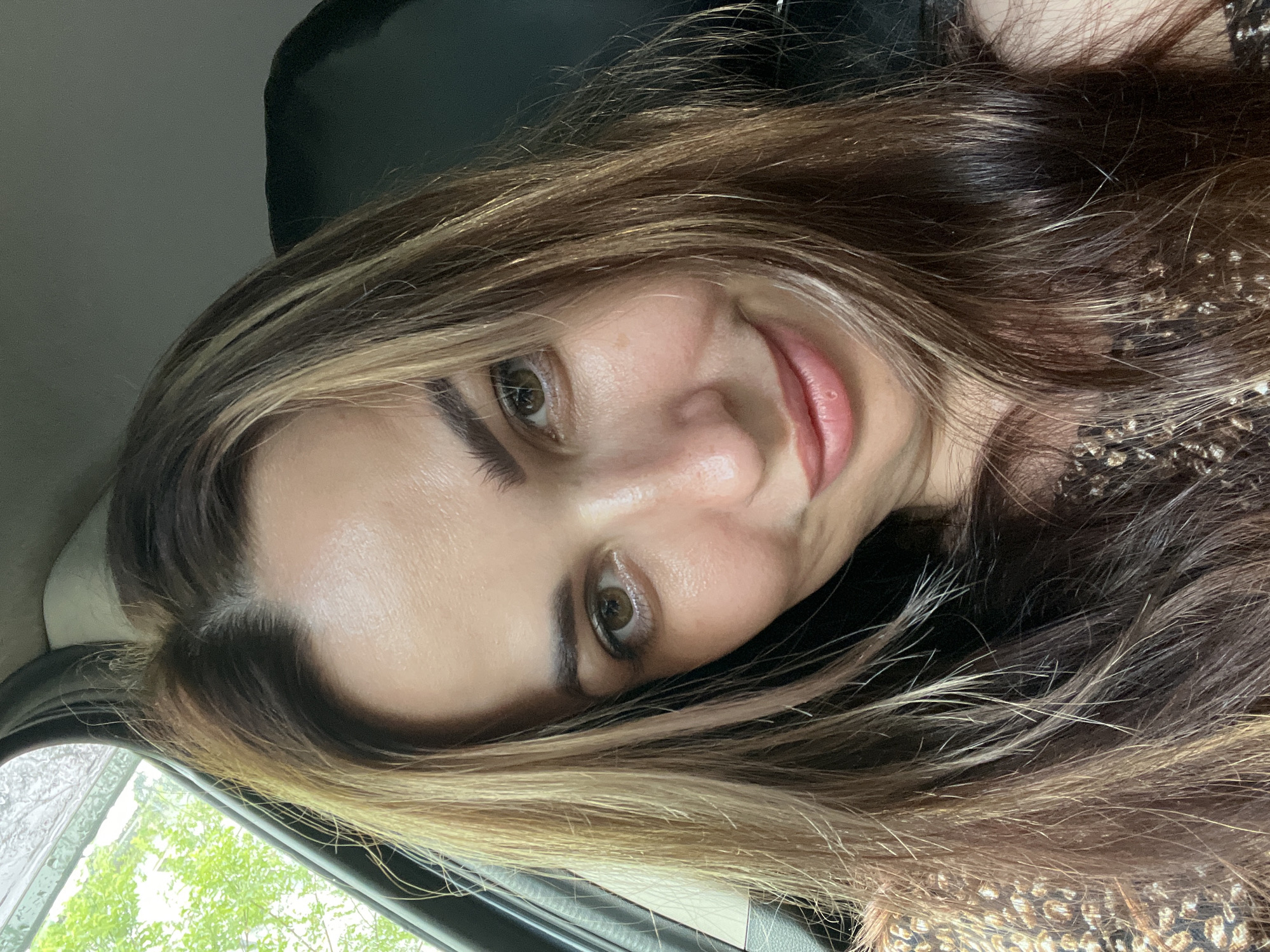 A medium shot captures a woman with long, layered brown hair with blonde highlights. She is sitting in the driver's seat of a vehicle, with the window on her left showing green foliage outside. Her head is tilted slightly to her right as she looks towards the camera, with a gentle smile on her face. She is wearing a dark, patterned top. The interior of the car, including a black headrest, is visible in the background.