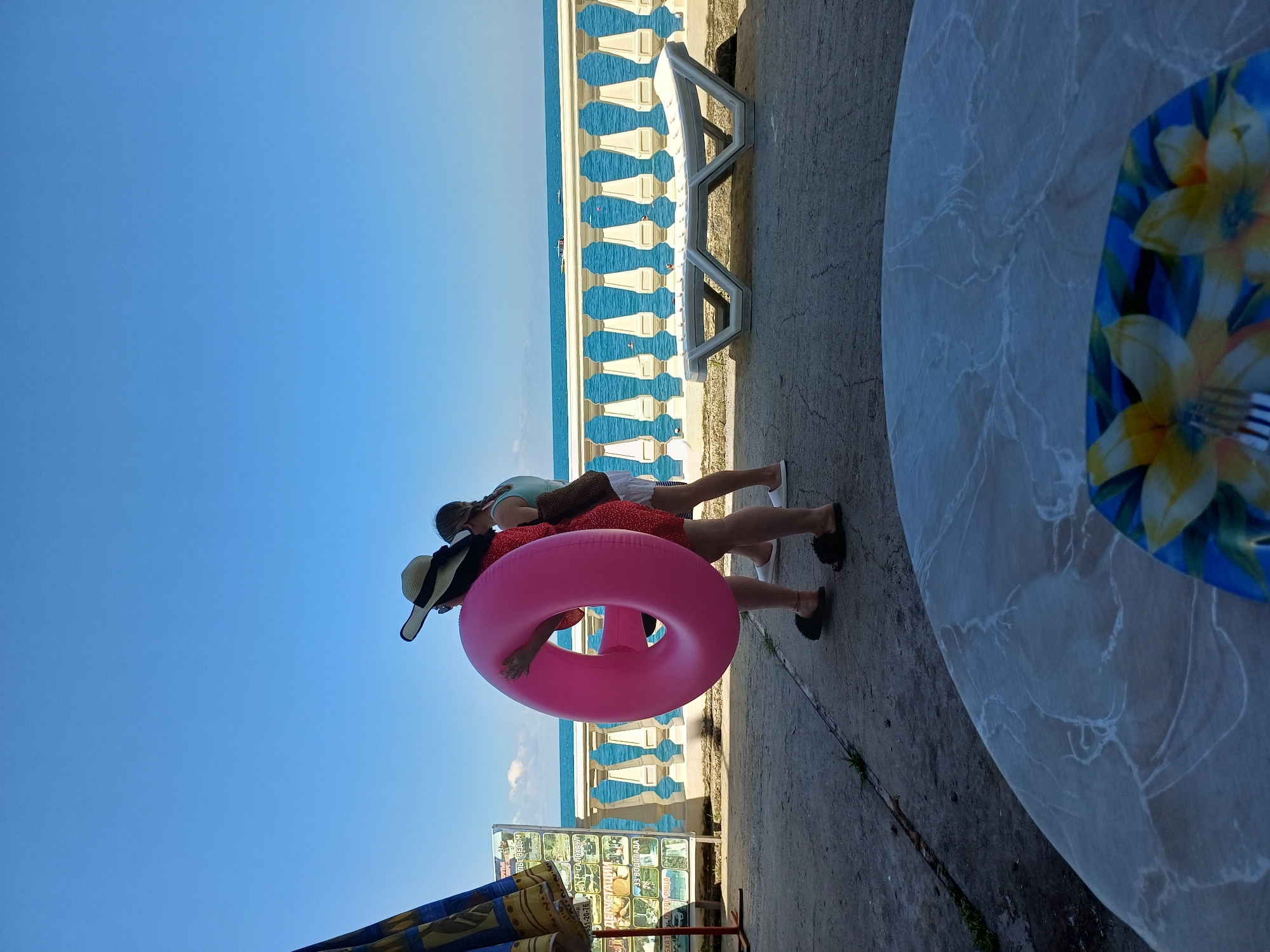 The image depicts two individuals walking on a concrete walkway near the Black Sea coast in Sochi, Russia. One person, wearing a wide-brimmed hat and a red polka-dot dress, carries a large pink inflatable ring. The other person, in a light blue top and white shorts, walks behind. In the background, a white balustrade lines the promenade overlooking the sea. A white plastic sun lounger is positioned on the concrete area adjacent to the balustrade. The sky is clear and blue. In the foreground, a round table with a marble-like surface is partially visible, with a plate decorated with yellow flowers and a fork resting on it. To the left, a portion of a blue and yellow striped beach umbrella is seen, along with a display of various advertisements or information boards. The overall scene suggests a sunny day at a seaside resort.