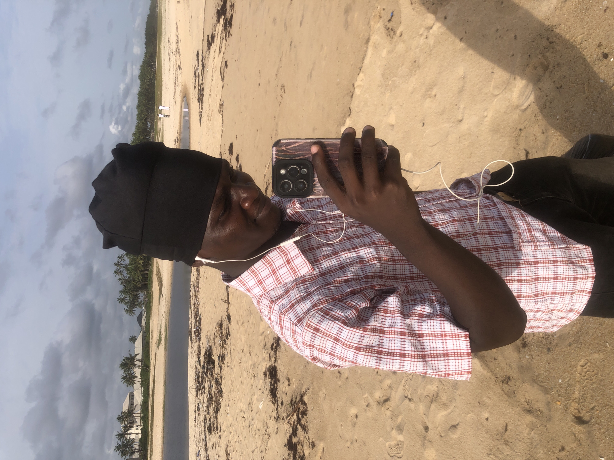 The image shows a person, presumably male, taking a selfie with a smartphone on a beach in Akodo, Nigeria. The person is wearing a black cap and a red-and-white checkered shirt and has earphones plugged in. The beach is sandy, with some seaweed or debris scattered around. In the background, there is a line of palm trees and buildings, and the sky is overcast. Two individuals can be seen on the horizon. The person appears to be standing still while taking the photo.