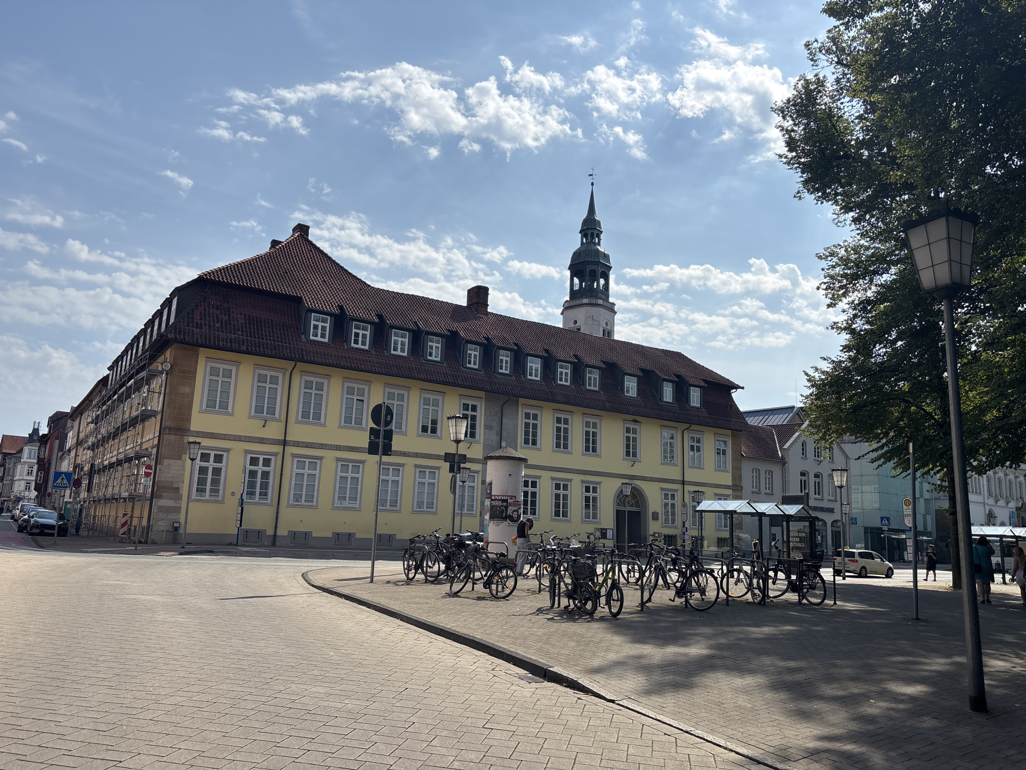 The image shows a street scene in Celle, Germany, on a sunny day with scattered clouds. In the foreground, a paved area with brick-like patterns leads to a street. To the left, a long, yellow building with a red tiled roof occupies a significant portion of the frame. This building features numerous windows with white frames on multiple floors. In the distance, a tall, dark green tower with a spire rises above the rooftops.
In front of the yellow building, a row of parked bicycles is visible, some of them in racks and others leaning against each other. A cylindrical structure, possibly a pillar with a poster attached, stands among the bicycles. A person is seen near this pillar, interacting with something on it.
Further down the street, to the right of the yellow building, other buildings of varying architectural styles are visible, including a white building and a structure with a glass facade. A bus stop shelter with a glass roof is present. A yellow taxi is driving away from the camera on the street. People are walking on the sidewalk near the bus stop.
To the right of the scene, a large tree with dense green foliage extends into the frame, and a street lamp with a square lantern is positioned in the foreground. The sky is blue with white, wispy clouds. The overall impression is a typical European city street with historical architecture and signs of everyday life, including transportation and pedestrian activity.