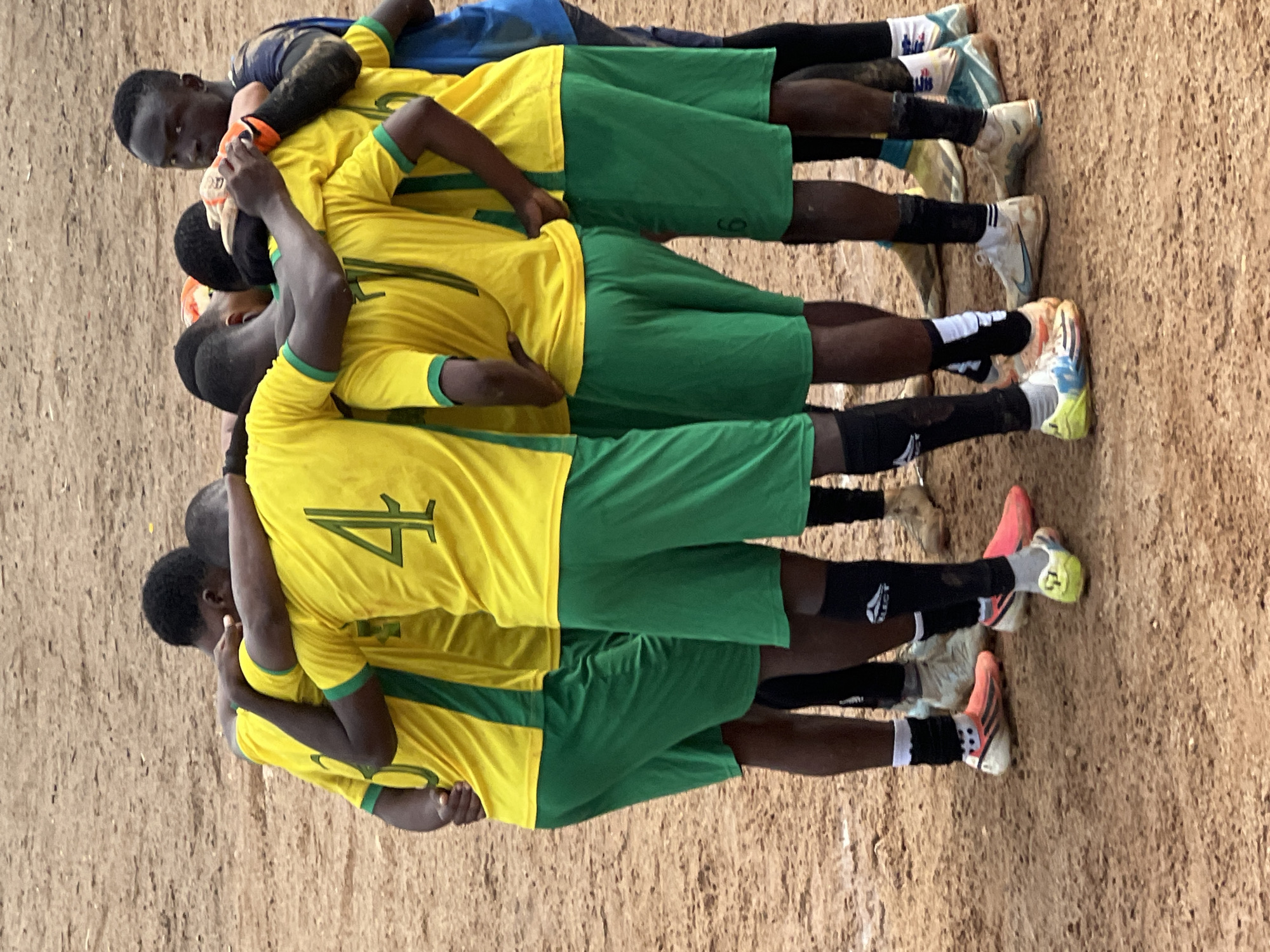 The image depicts a group of young male soccer players in a huddle on a dirt field. They are wearing yellow jerseys with green accents and green shorts. Several players have numbers visible on their jerseys, including a "4" and a "7." The players are embracing each other, appearing to be strategizing or celebrating. The ground is a dusty, reddish-brown dirt, indicative of a typical playing surface in many parts of Jos, Nigeria, especially in less developed areas or local fields. The background is blurred, suggesting a focus on the players. The overall atmosphere is one of camaraderie and focus on the sport.