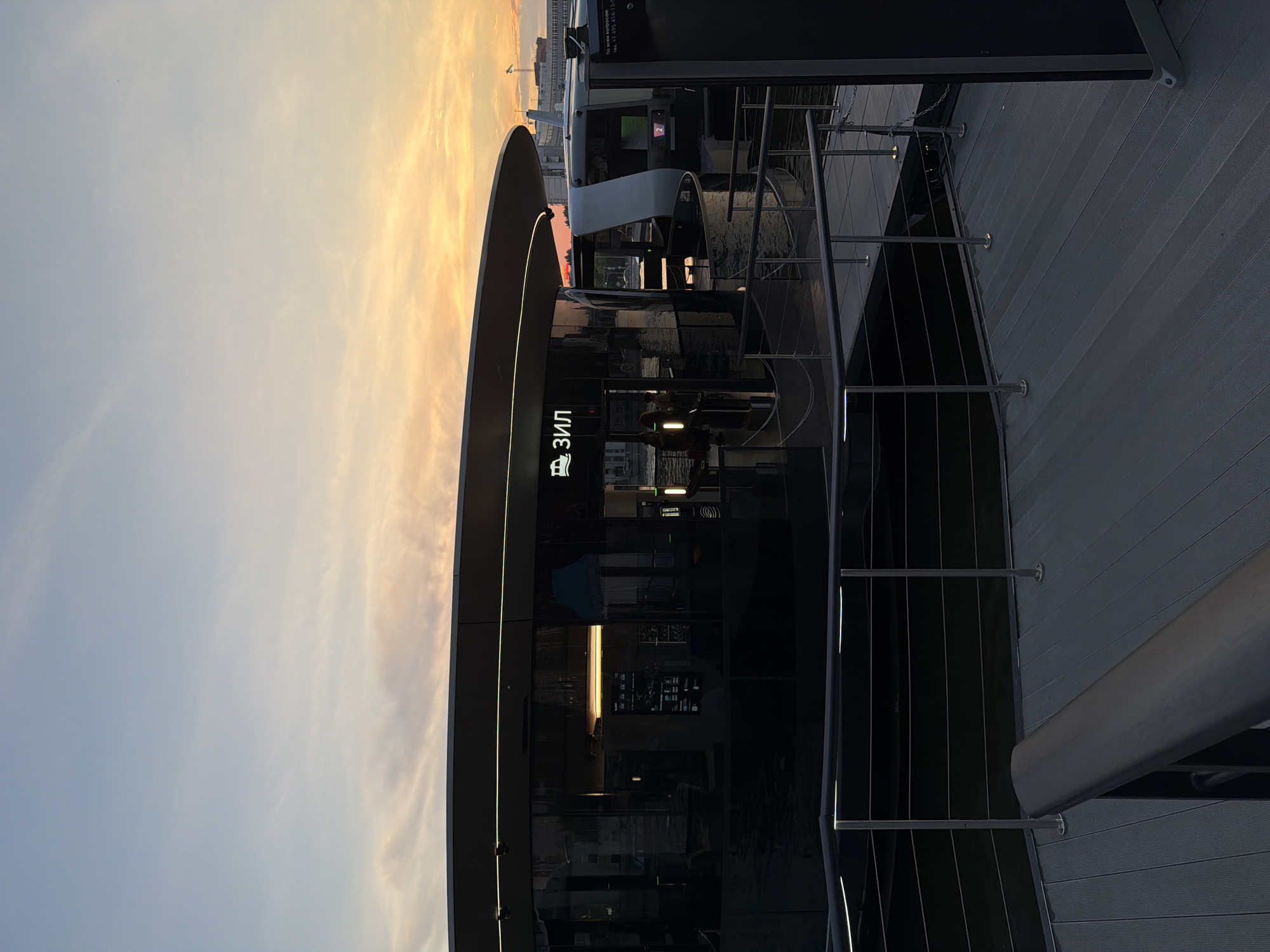 This is a photograph of a modern, sleek building situated on the water, likely a pier or a floating structure. The building features a curved, dark facade with extensive glass windows, reflecting the twilight sky and surrounding lights. A prominent sign with Cyrillic lettering that reads "ЗИЛ" (ZIL) is illuminated above the entrance, accompanied by a small icon of a boat or ferry. The sky above displays a gradient of soft blues and oranges, characteristic of a sunset.
The building appears to be part of a transportation hub, as indicated by the boat icon and a nearby vessel that resembles a modern passenger ferry or water taxi. There are people visible inside the building, silhouetted against the interior lighting, suggesting it is operational. A metal railing and a wooden deck structure are in the foreground, providing access to the building. The overall impression is one of contemporary urban infrastructure set against a tranquil, atmospheric sky.
The location is identified as Moscow, Russia, specifically the ZIL pier on the Moskva River, part of the city's expanding water transport network.